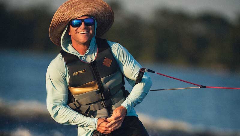 Shaun Murray wakeboarding on water, wearing a wide-brimmed hat, sunglasses, a life vest, and shorts. The person is smiling and holding a rope. The water splashes around them under a clear sky.