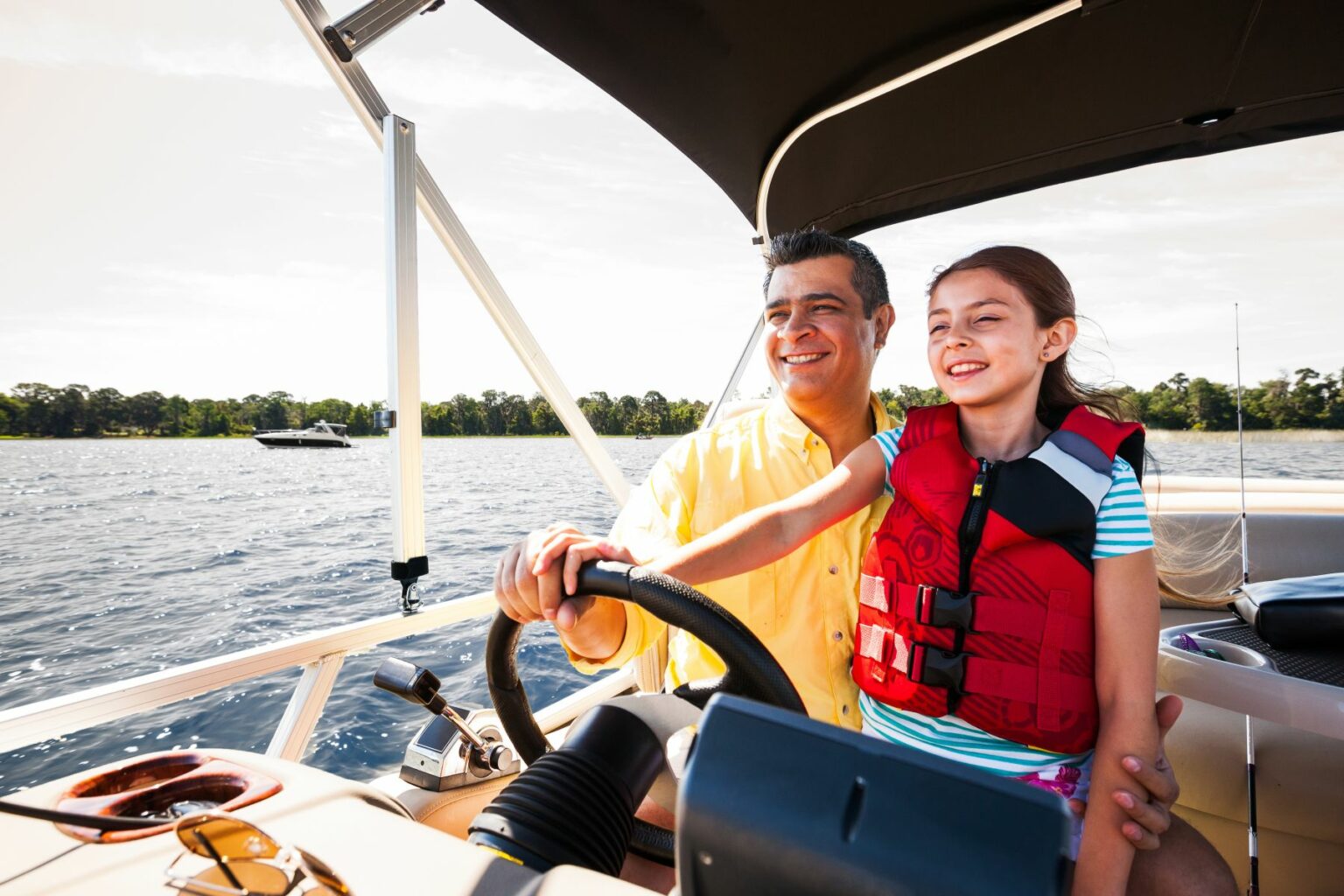 Royalty-free image - A man and a young girl are smiling while steering a boat. The girl is wearing a red life jacket, and they are surrounded by water with a tree-lined shore in the background under a clear sky.