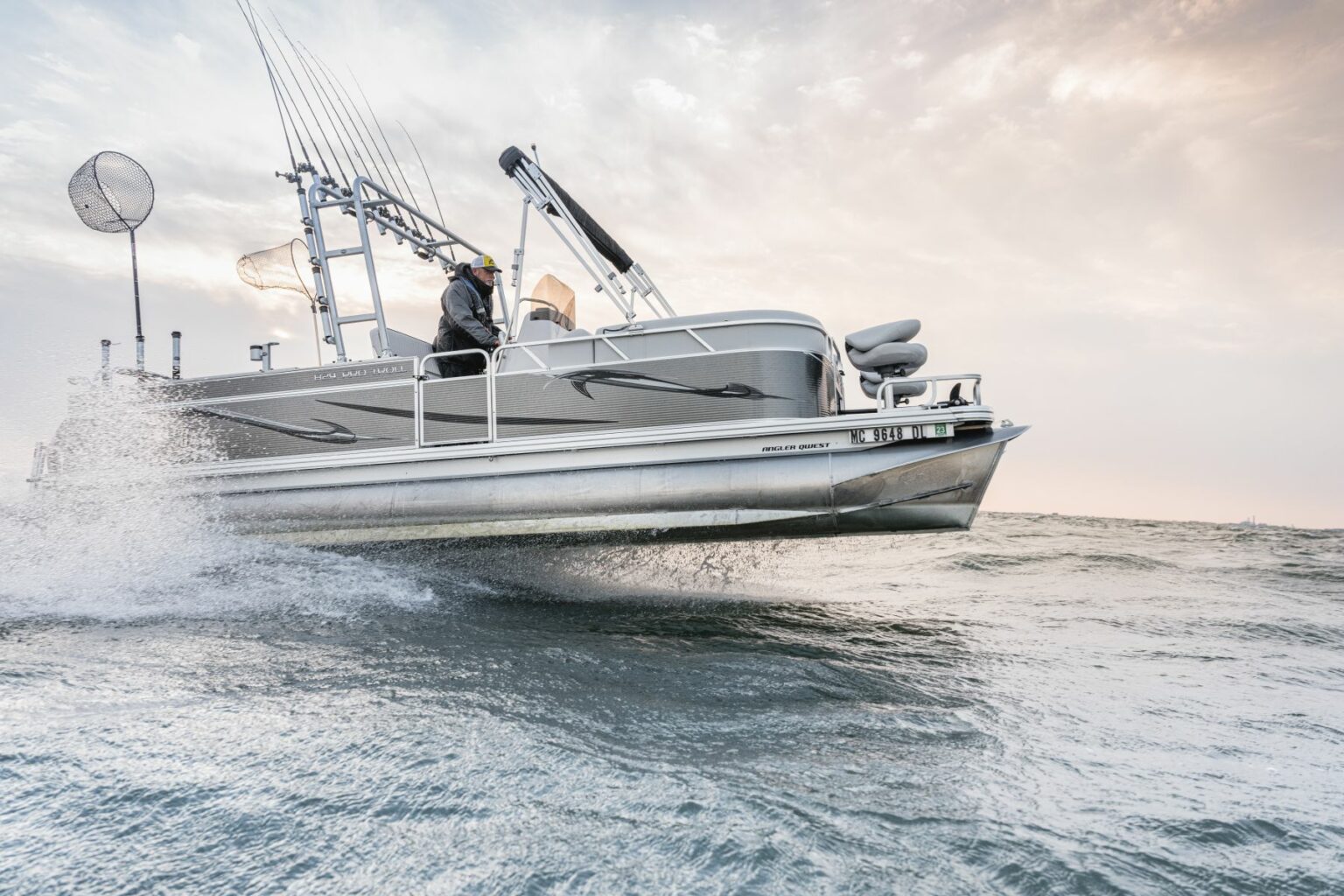 Royalty-free image - A silver fishing boat speeds across choppy waters, creating a splash. The boat is equipped with numerous fishing rods and a net. The sky is overcast, providing a dramatic backdrop to the scene.