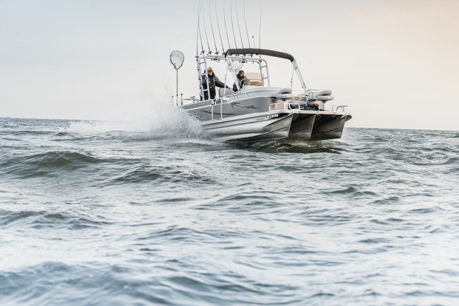 Royalty-free image - A small fishing boat speeds across choppy waters with two people on board. Several fishing rods are mounted on the boat, and one person holds a fishing net. The sky is overcast, giving the scene a dynamic and adventurous feel.