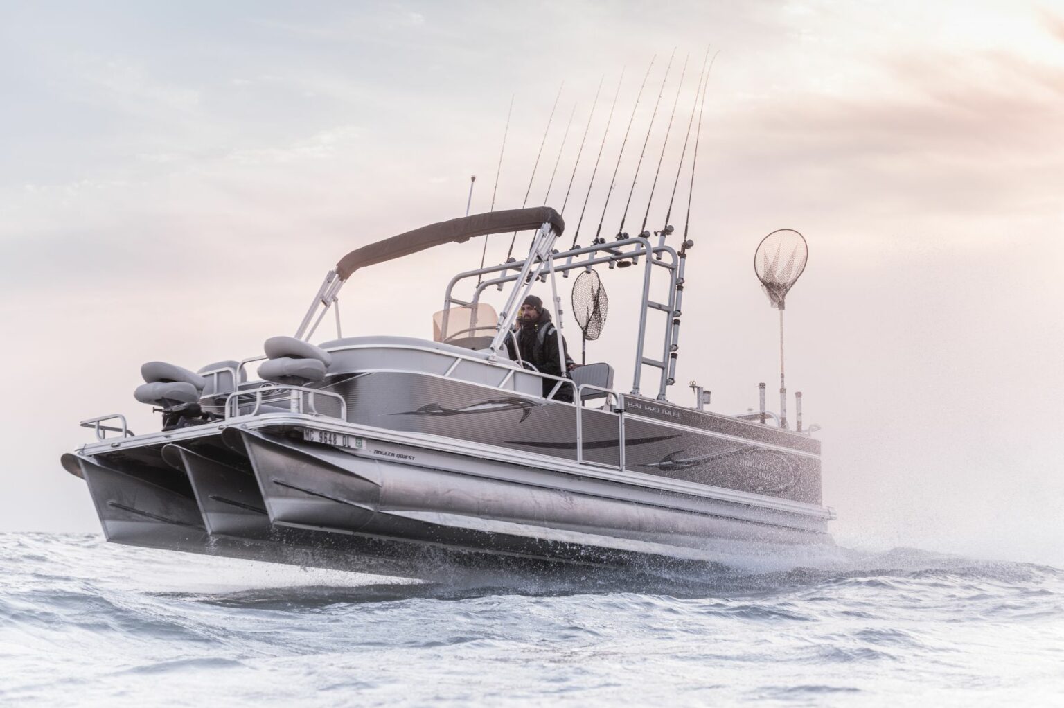 Royalty-free image - A sleek, modern pontoon boat with multiple fishing rods and nets mounted on the back moves swiftly over the ocean. The sky is overcast, lending a soft and muted atmosphere to the scene.