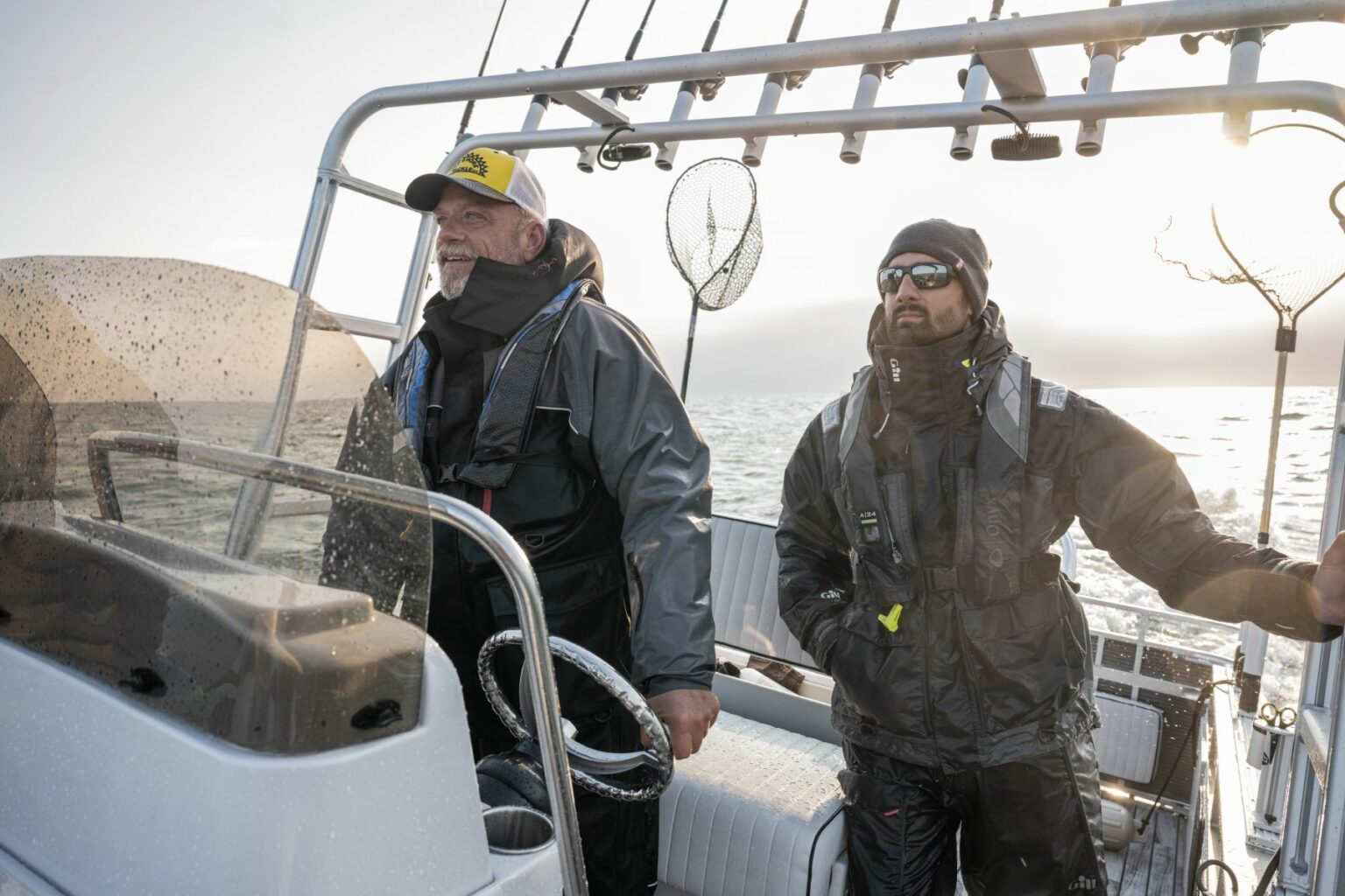 Royalty-free image - Two people in waterproof gear are on a boat in the open sea. One person steers while the other stands beside, both looking out. The sun shines through the cloudy sky, and fishing equipment is mounted on the boat.