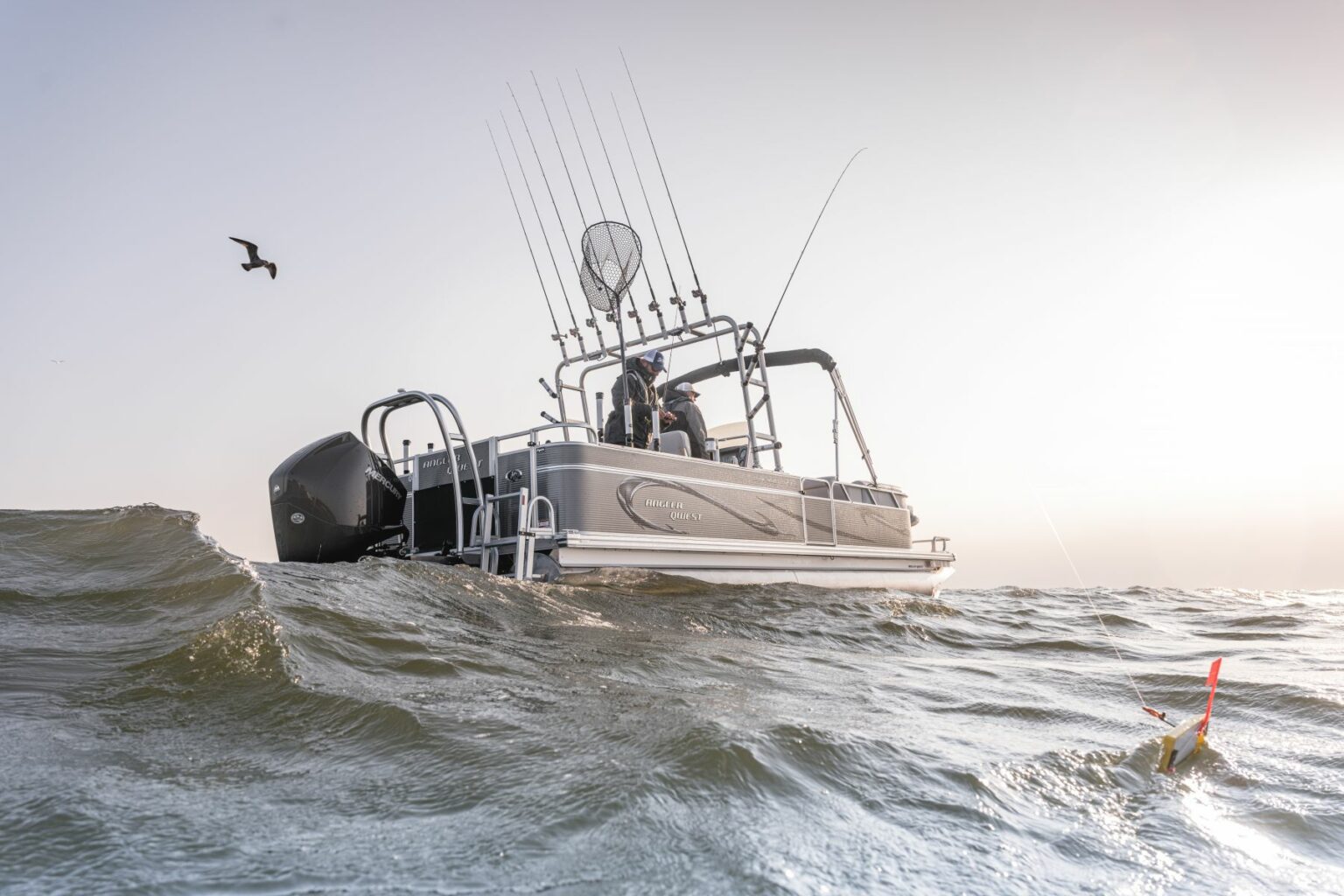 Royalty-free image - A boat with fishing rods and a net is navigating ocean waves under a clear sky. A bird flies nearby, and a fishing lure floats on the water in the foreground. The scene is serene and captures a moment of sport fishing adventure.