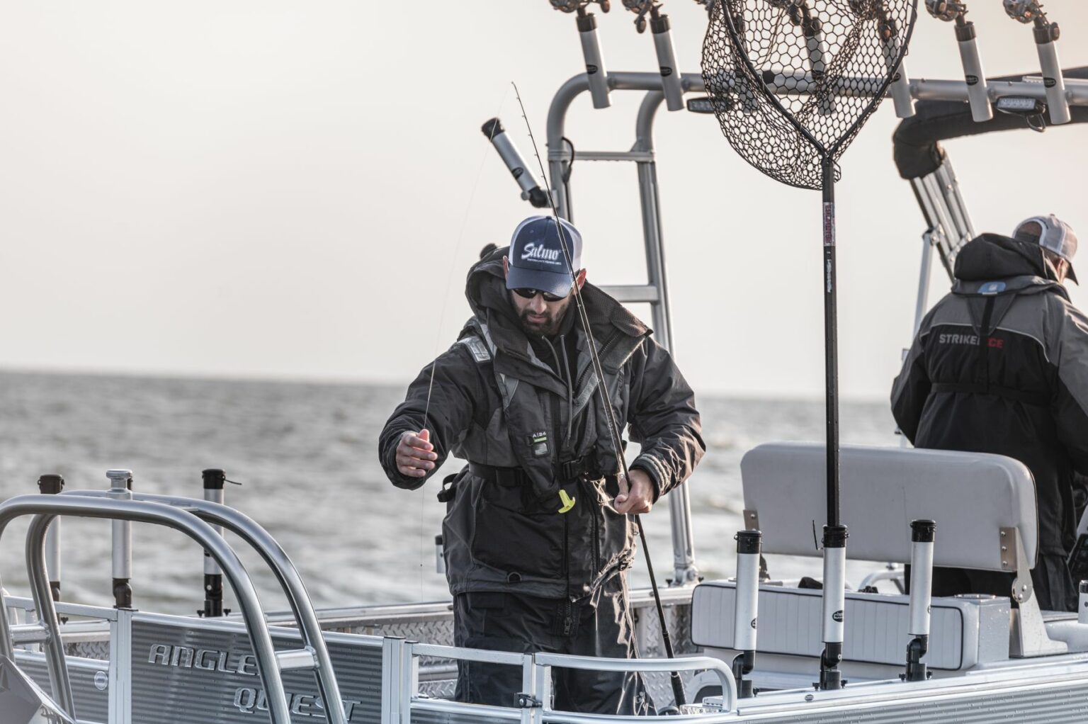 Royalty-free image - A person in a jacket and cap is standing on a boat, holding fishing equipment. The boat is on a body of water, and fishing nets and rods are visible nearby. Another person is in the background, also on the boat.