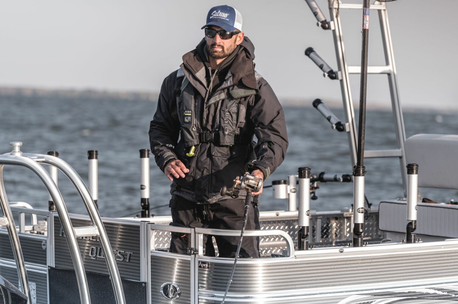 Royalty-free image - A person wearing a dark jacket and cap is standing on a boat, holding a fishing rod. The boat is equipped with metal railings and the background shows a body of water and a partly cloudy sky.