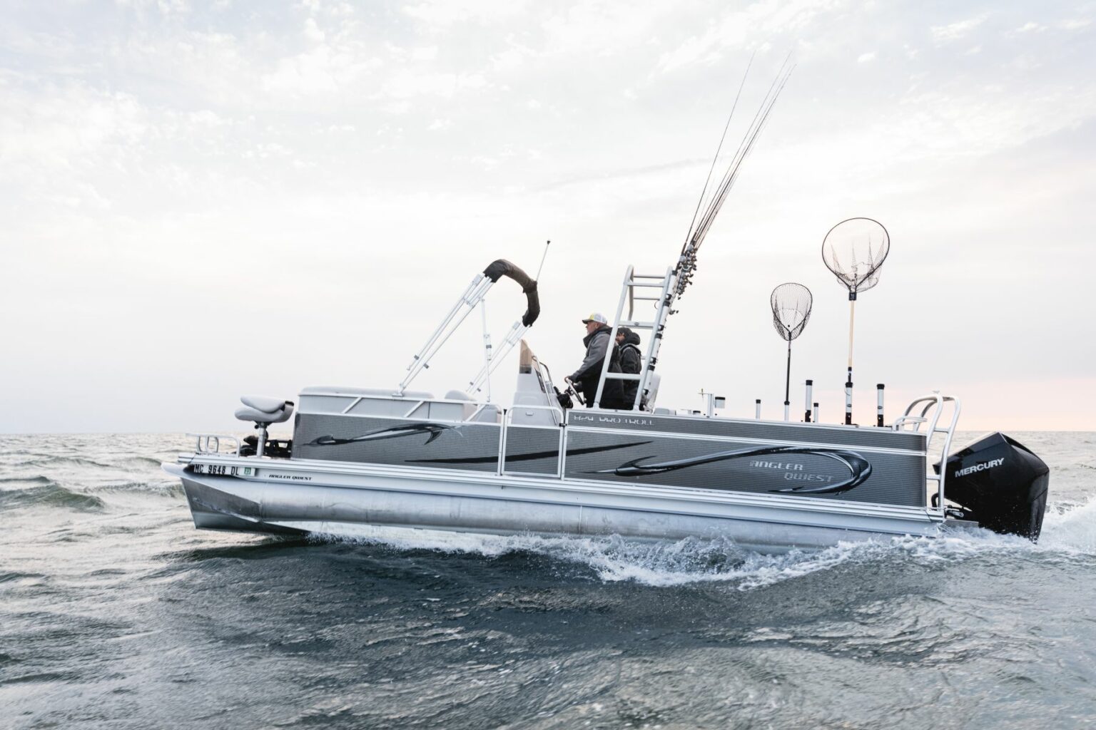 Royalty-free image - A man steers a gray fishing boat with several fishing rods and nets secured on board. The boat moves across a calm, expansive body of water under a cloudy sky.