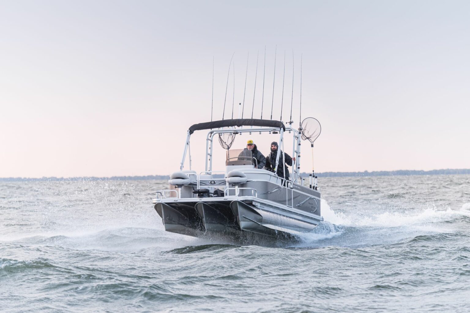 Royalty-free image - A fishing boat with multiple fishing rods moves quickly across a body of water, creating a wake. Two people are visible on the boat, and the sky is clear with a distant shoreline in the background.