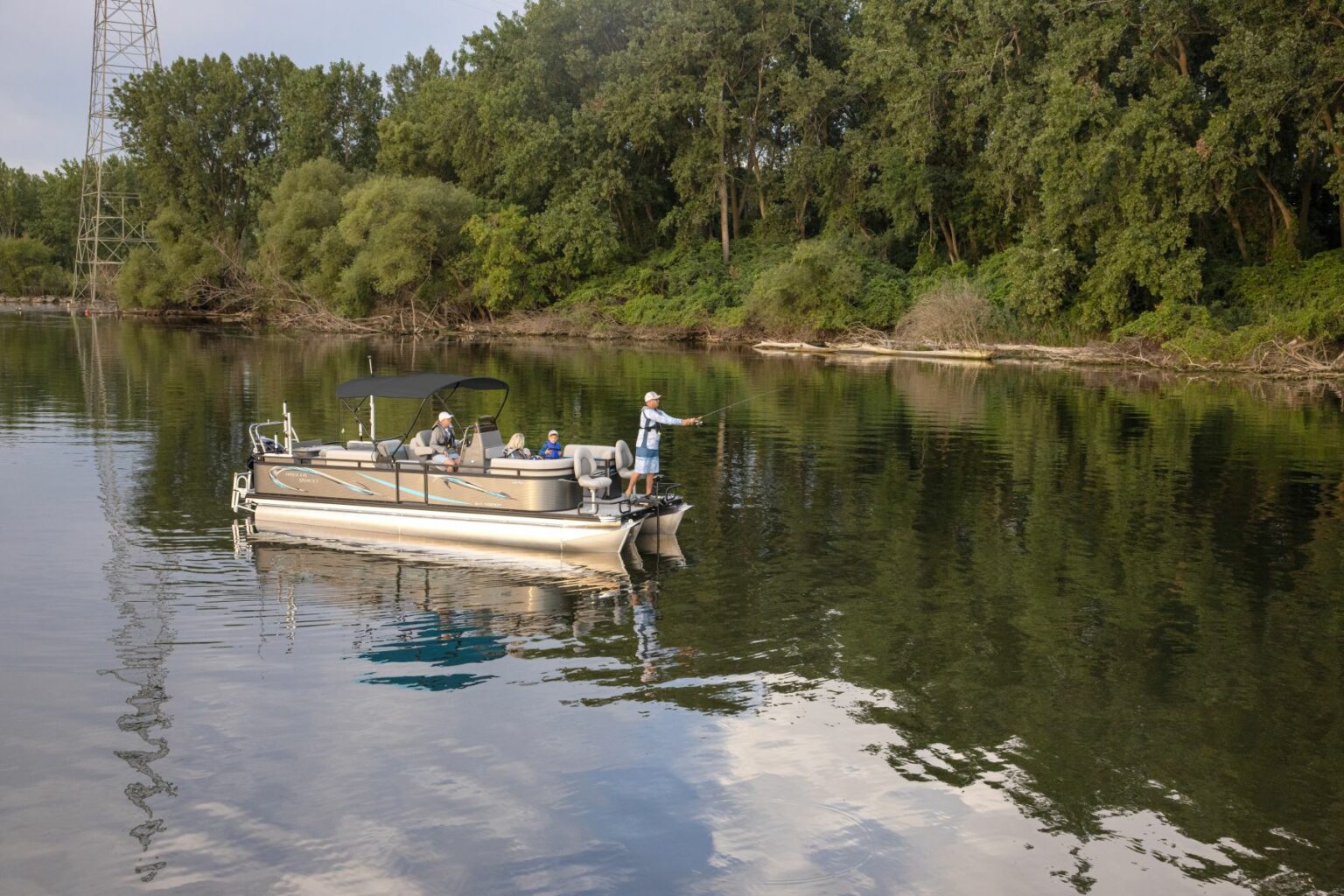 Royalty-free image - A group of people enjoys an outing on a pontoon boat on a calm river. One person is fishing while others relax under the boat&#039;s canopy. The surrounding landscape features dense foliage and tall trees reflecting in the water.