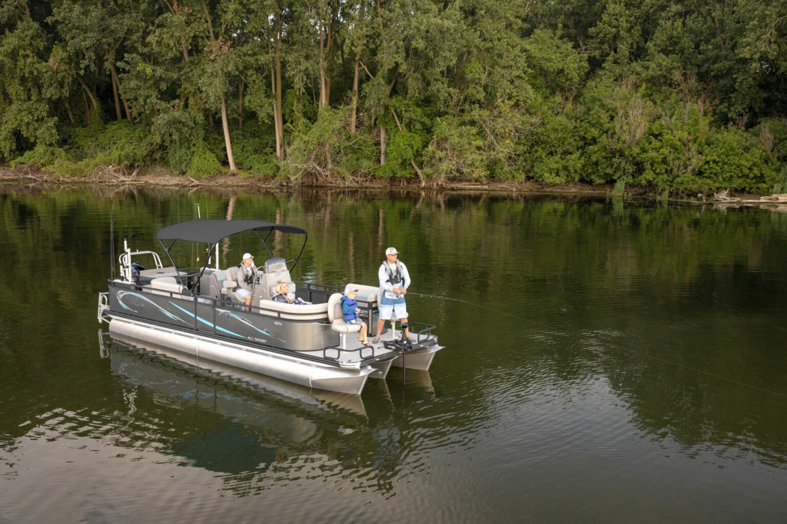 Royalty-free image - A group of people are on a pontoon boat in a calm river. The boat is near a forested shoreline with lush green trees. Two individuals are engaged in fishing, while the others are seated, enjoying the scenic surroundings.