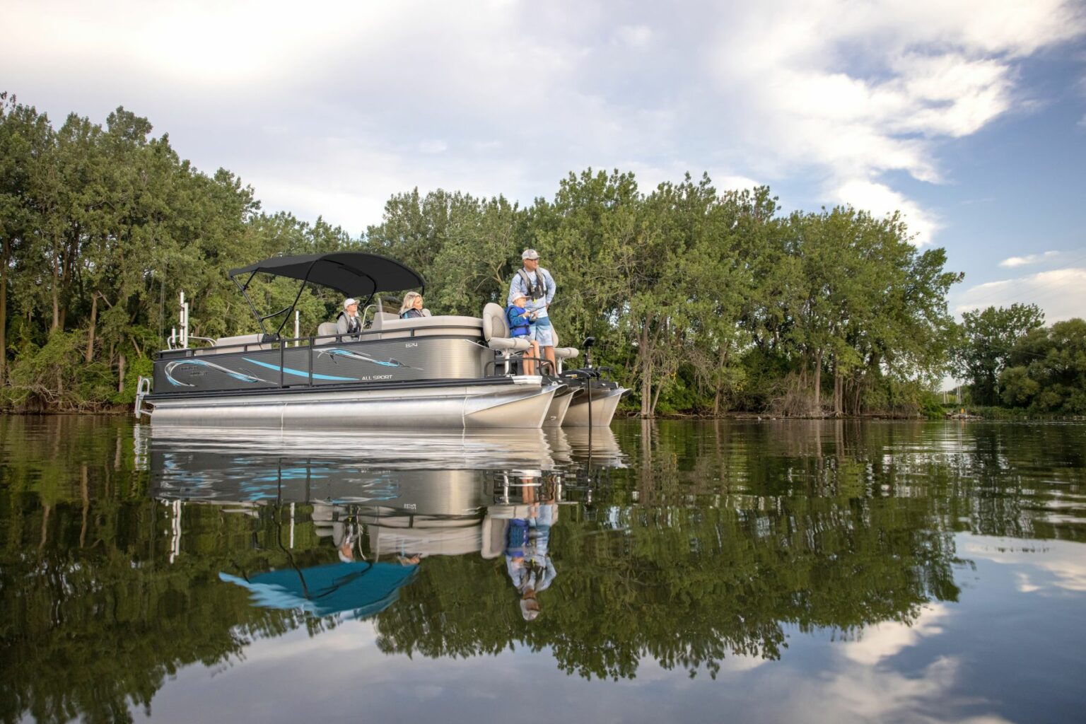 Royalty-free image - A family is fishing on a pontoon boat in a calm, tree-lined lake. The sky is partly cloudy, and the water reflects the boat and trees. Some people are sitting, and one person is standing with a fishing rod.