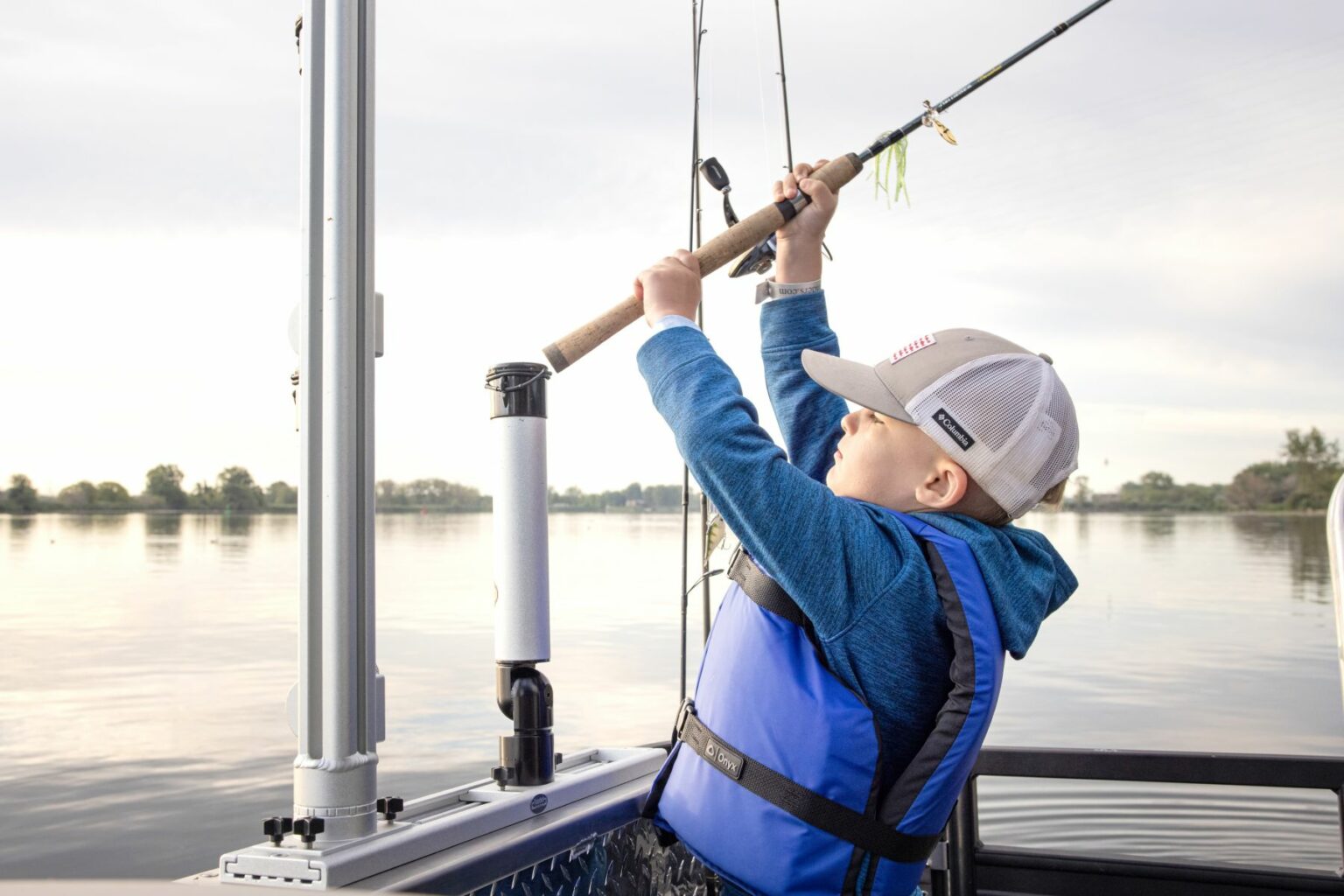 Royalty-free image - A young child wearing a USCG-approved blue life jacket and a baseball cap is fishing from a boat. The child holds a fishing rod, gazing up at the line. In the background, a calm lake with trees lines the shoreline.