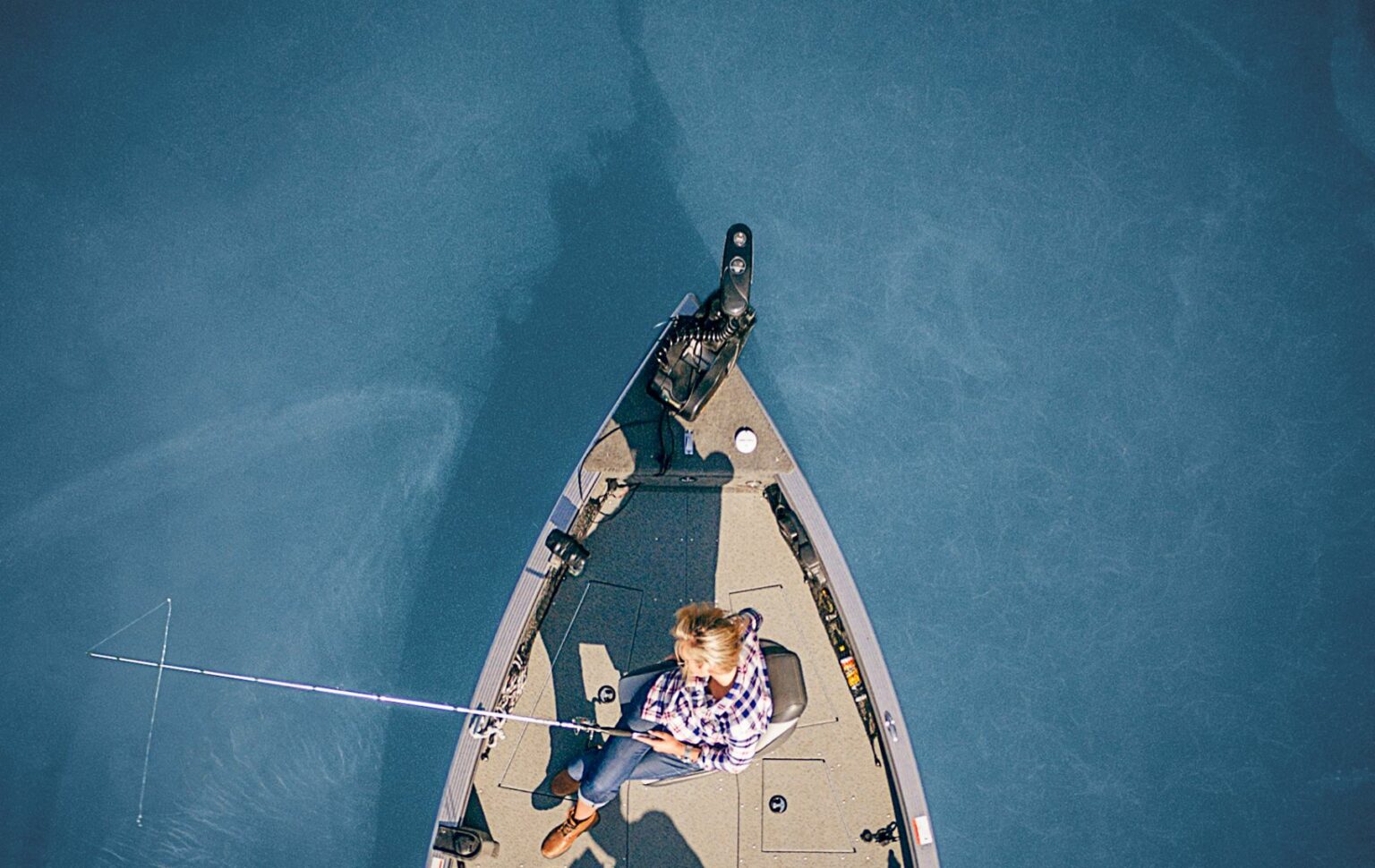 Royalty-free image - A person in a checkered shirt is sitting in a small jon boat, holding a fishing rod. The boat glides on a calm, blue body of water, with sunlight casting enchanting shadows of both the boat and angler on the water&#039;s surface.