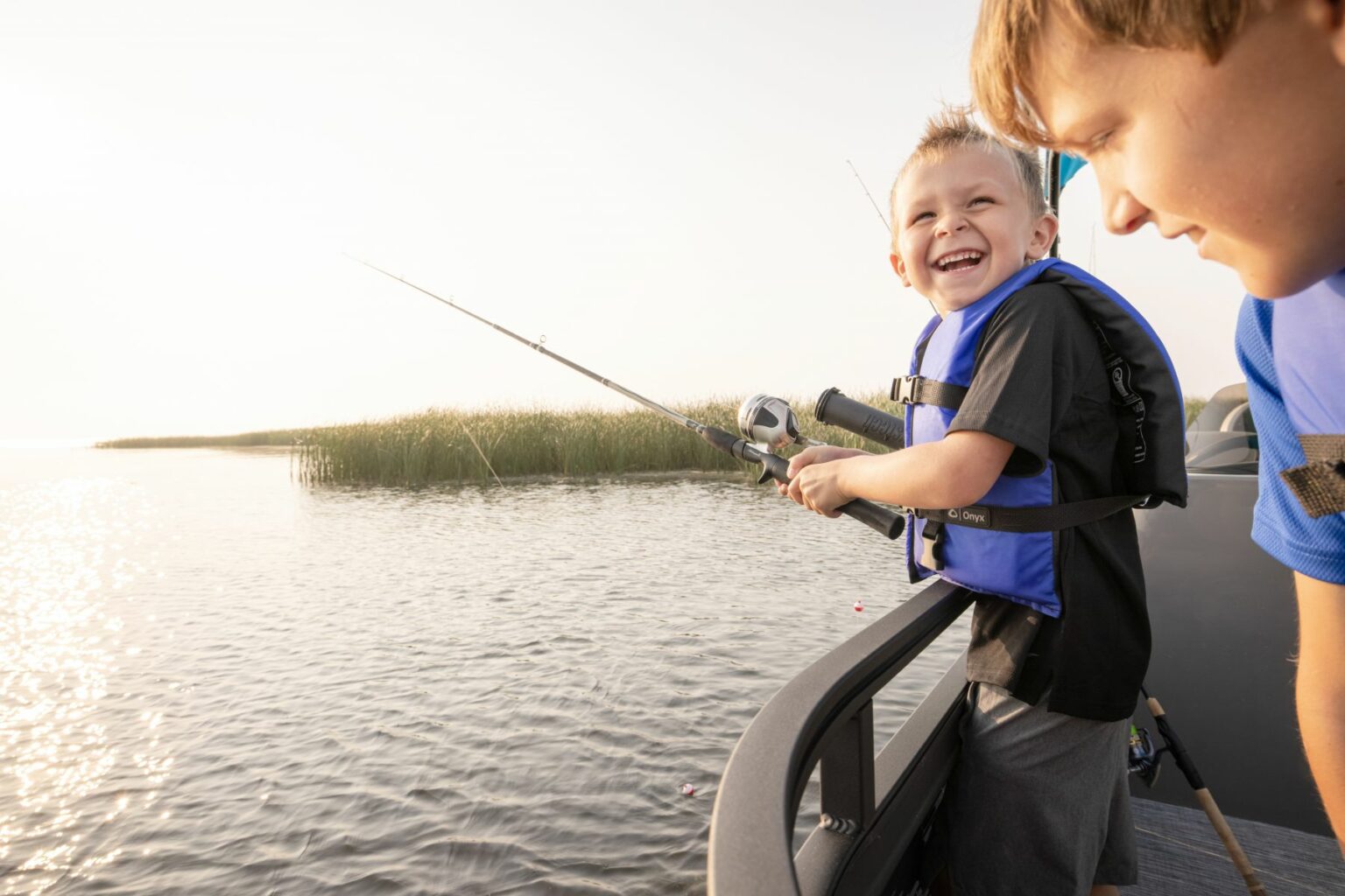 Royalty-free image - A young child wearing a USCG-approved life jacket and holding a fishing rod smiles joyfully while fishing from a boat. An adult nearby looks on as the sun sets over the calm lake with reeds in the background.