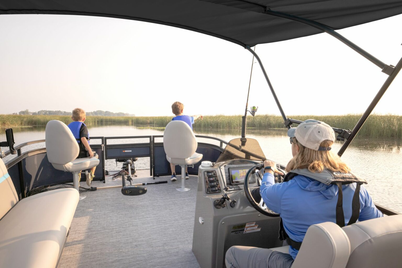 Royalty-free image - A person steers a boat along a calm, narrow waterway bordered by tall grass. Two children sit at the front of the boat, looking out at the water. The sun is low, casting a soft light over the scene.