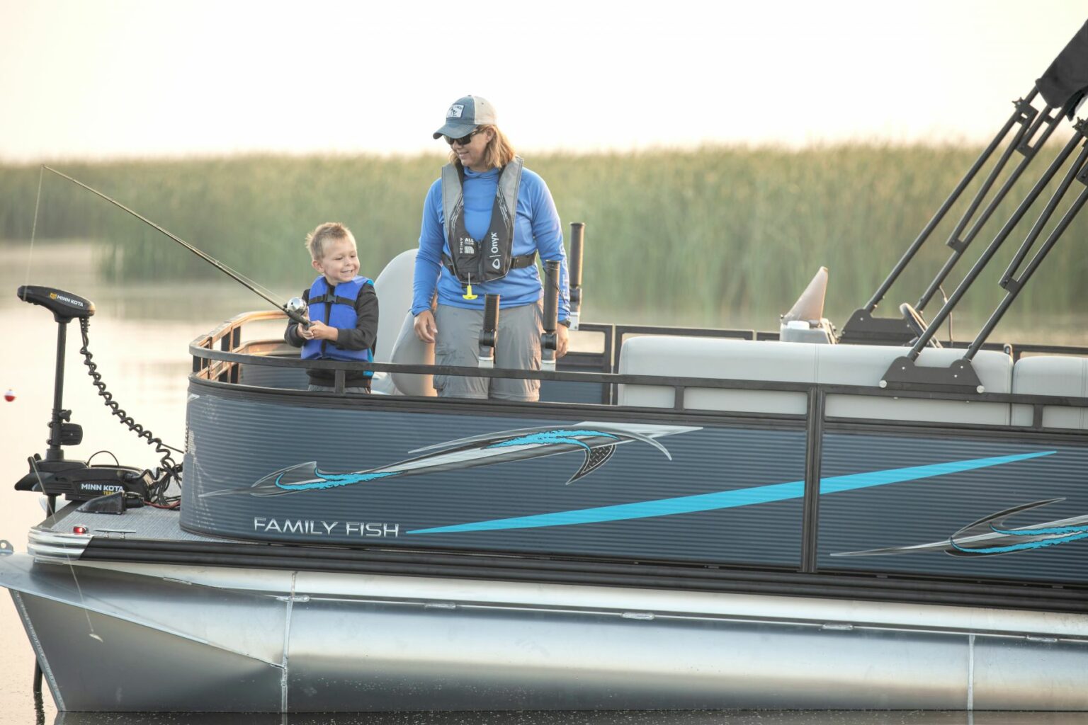 Royalty-free image - A woman and a young child are on a pontoon boat, both wearing life jackets. The child is holding a fishing rod, smiling at the woman. They are surrounded by calm water and reeds in the background. The boat has &quot;Family Fish&quot; written on the side.