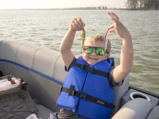 License-free image A child in a blue life jacket and reflective sunglasses is standing on a boat, holding up two small fish lures proudly. The boat is on a calm lake with a tree-lined shore visible in the background. A tackle box is seen at the foreground of the boat.