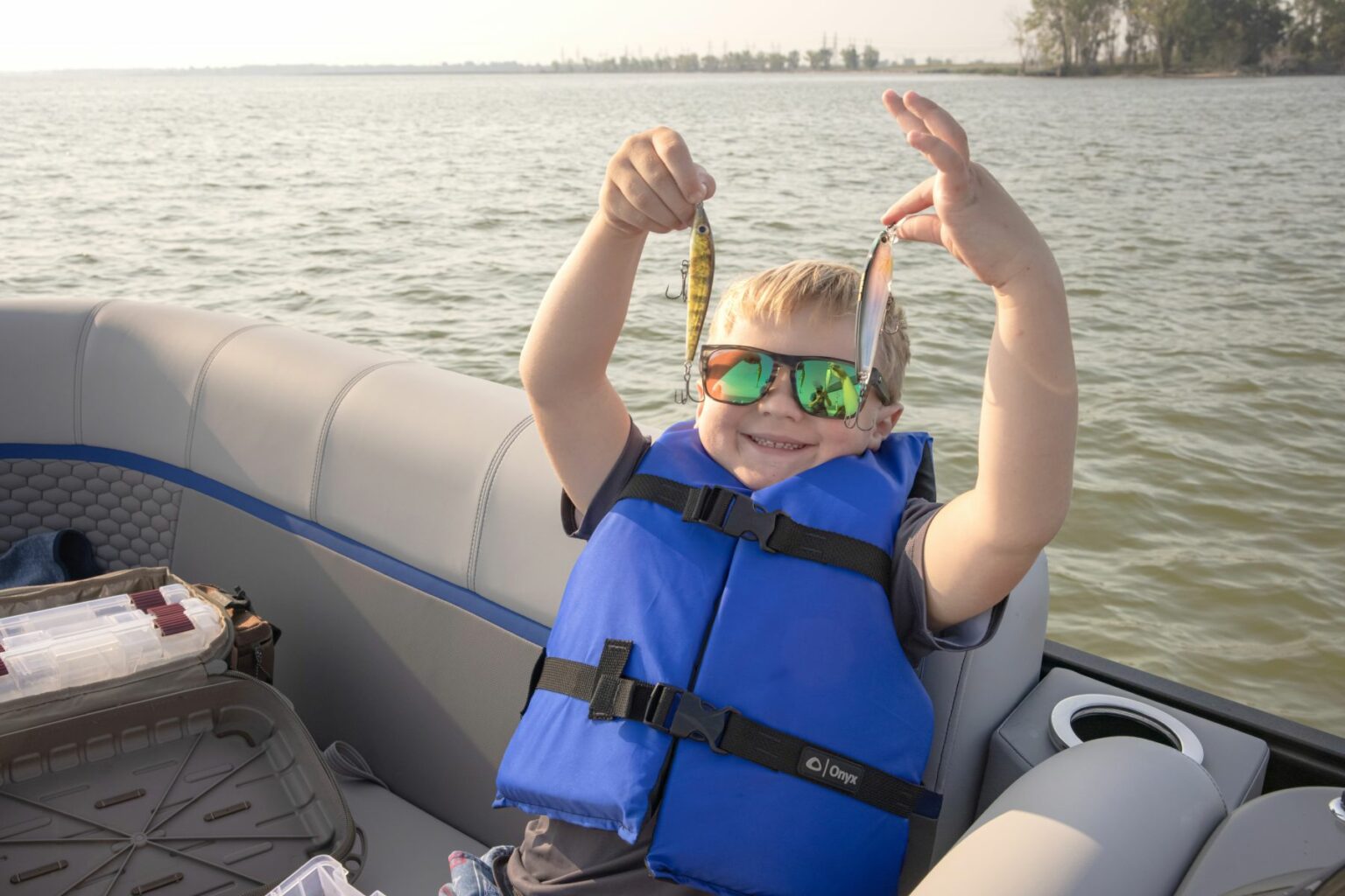 License-free image A child in a blue life jacket and reflective sunglasses is standing on a boat, holding up two small fish lures proudly. The boat is on a calm lake with a tree-lined shore visible in the background. A tackle box is seen at the foreground of the boat.