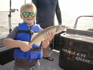 License-free image A child in a blue life jacket and sunglasses smiles while holding a fish on a boat. Another person stands nearby, partially visible. The sun is shining, and the boat&#039;s equipment and surroundings suggest they are out on a fishing trip.
