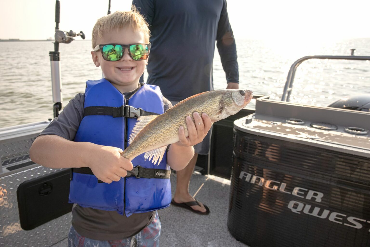 License-free image A child in a blue life jacket and sunglasses smiles while holding a fish on a boat. Another person stands nearby, partially visible. The sun is shining, and the boat&#039;s equipment and surroundings suggest they are out on a fishing trip.