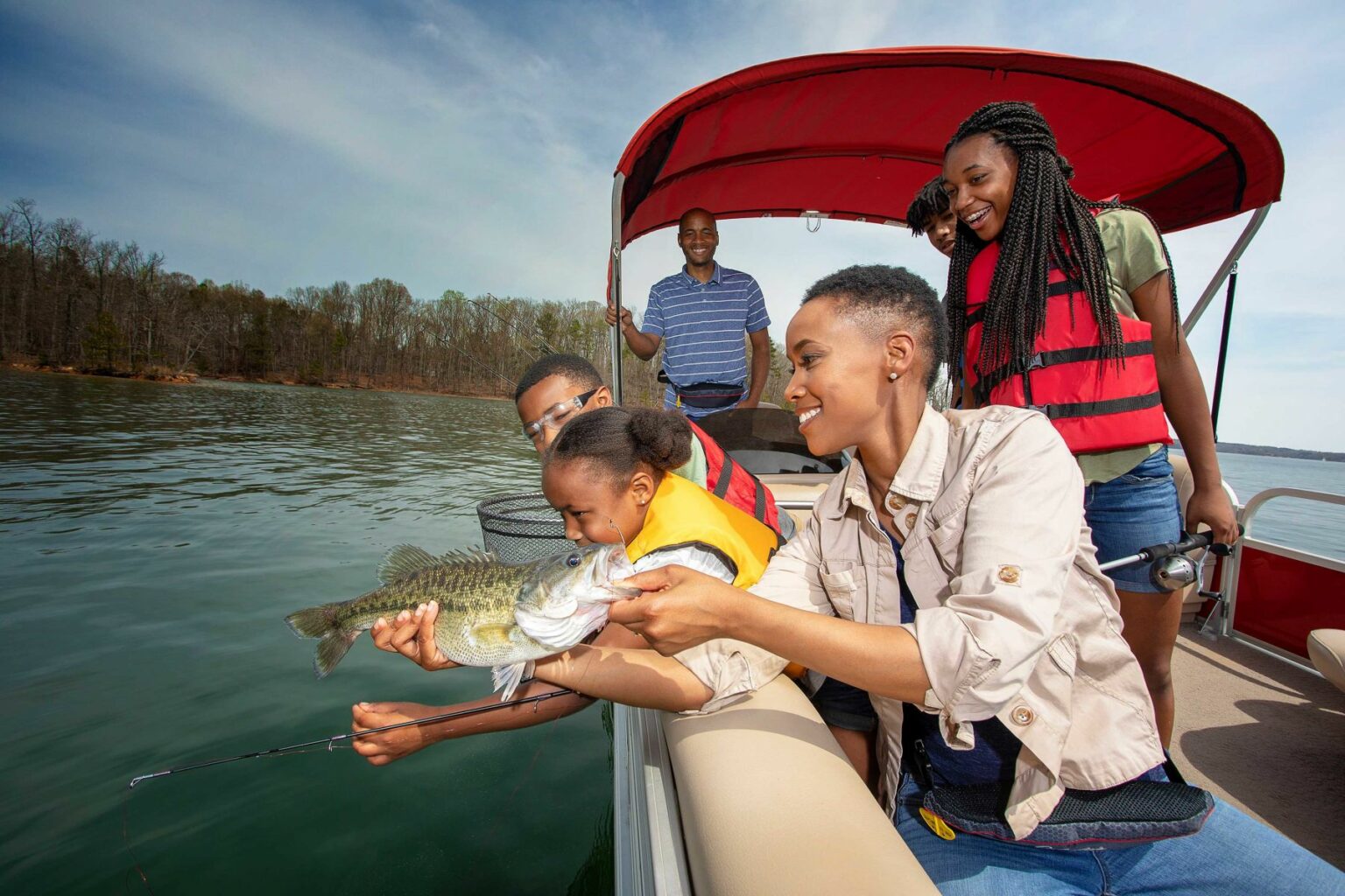 Royalty-free image - A family of five on a boat, smiling as they catch a large fish. Everyone is wearing life jackets. The water and forested shoreline are visible in the background. The mood is cheerful and lively.