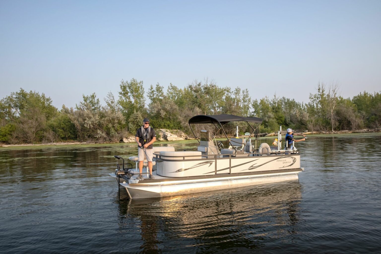 License-free image -Two people fishing from a pontoon boat on a calm lake. One person stands, casting a line, while the other sits near the back. The boat is equipped with a canopy, and trees line the distant shore under a clear blue sky.