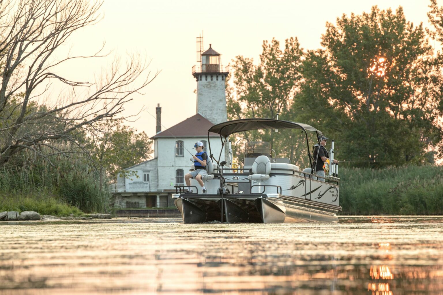 License-free image A pontoon boat with two passengers in lifevests cruises on a calm body of water at sunset, heading towards a historic white lighthouse standing among trees and tall grass on the shore. The sky is tinged with warm, golden hues.