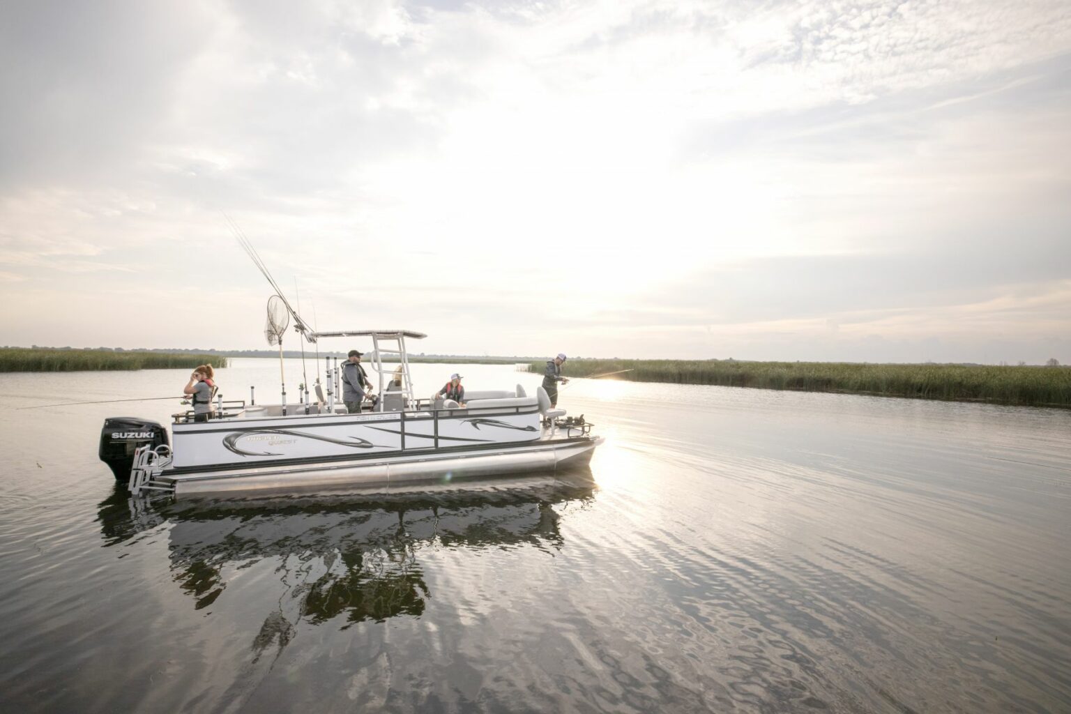 Royalty-free image - A small group of people are on a pontoon boat in a calm, expansive body of water. The sun is low, creating a bright glare on the water&#039;s surface. The surrounding landscape features marshy vegetation and an overcast sky.