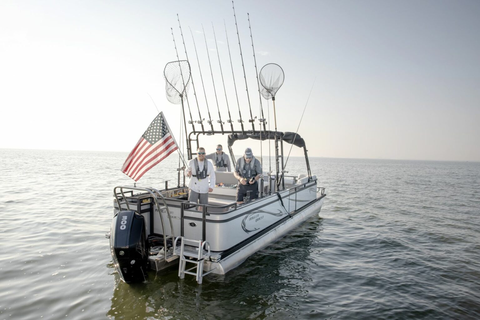 Royalty-free image - A fishing boat on calm waters with three people onboard. They are standing near fishing rods and a U.S. flag is mounted at the back. Two fishing nets are visible above them against a clear sky.