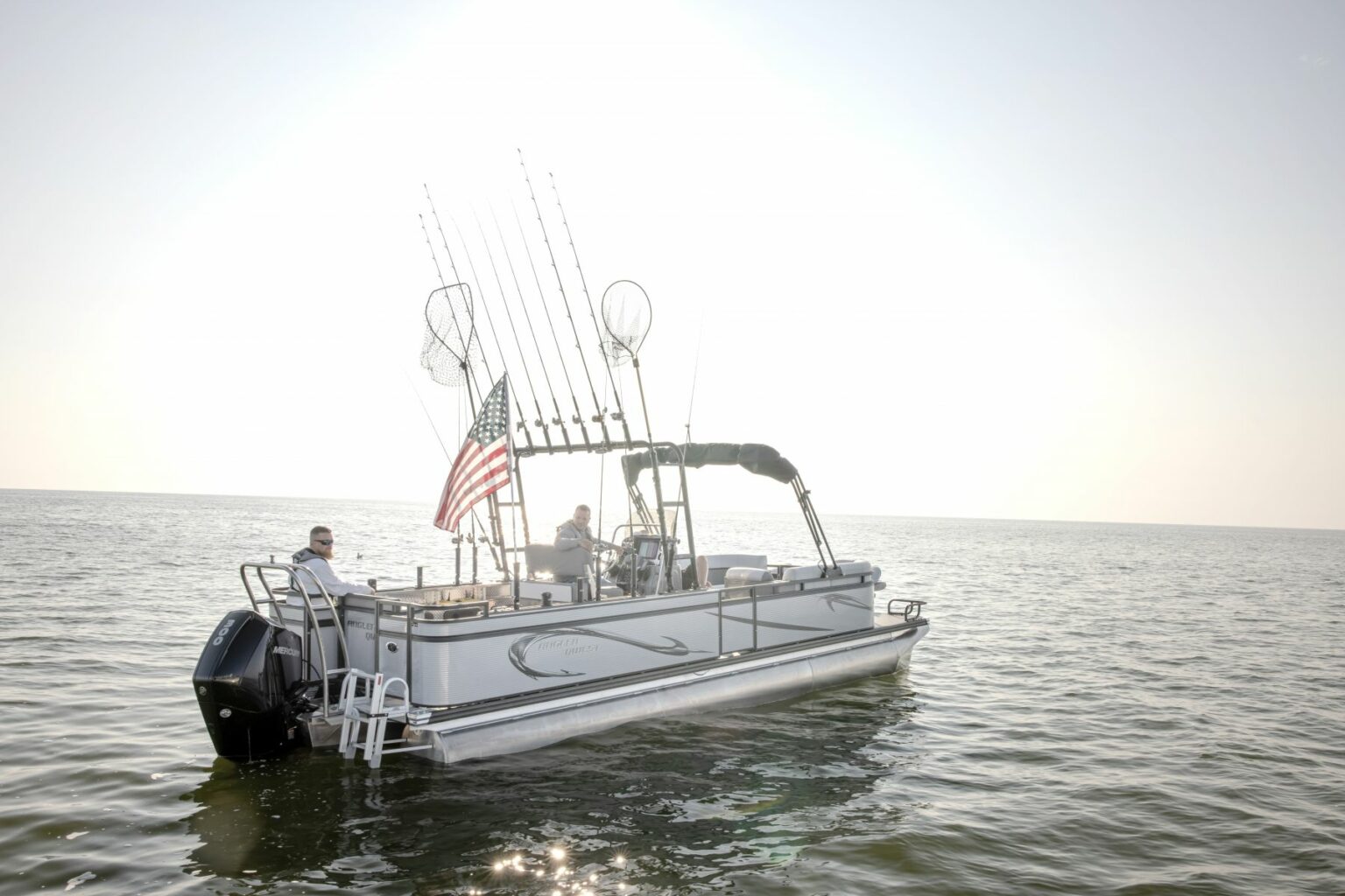 Royalty-free image - A pontoon boat with fishing rods and an American flag is floating on a calm lake under a bright sky. Two people are on the boat; one is sitting at the back while the other steers.