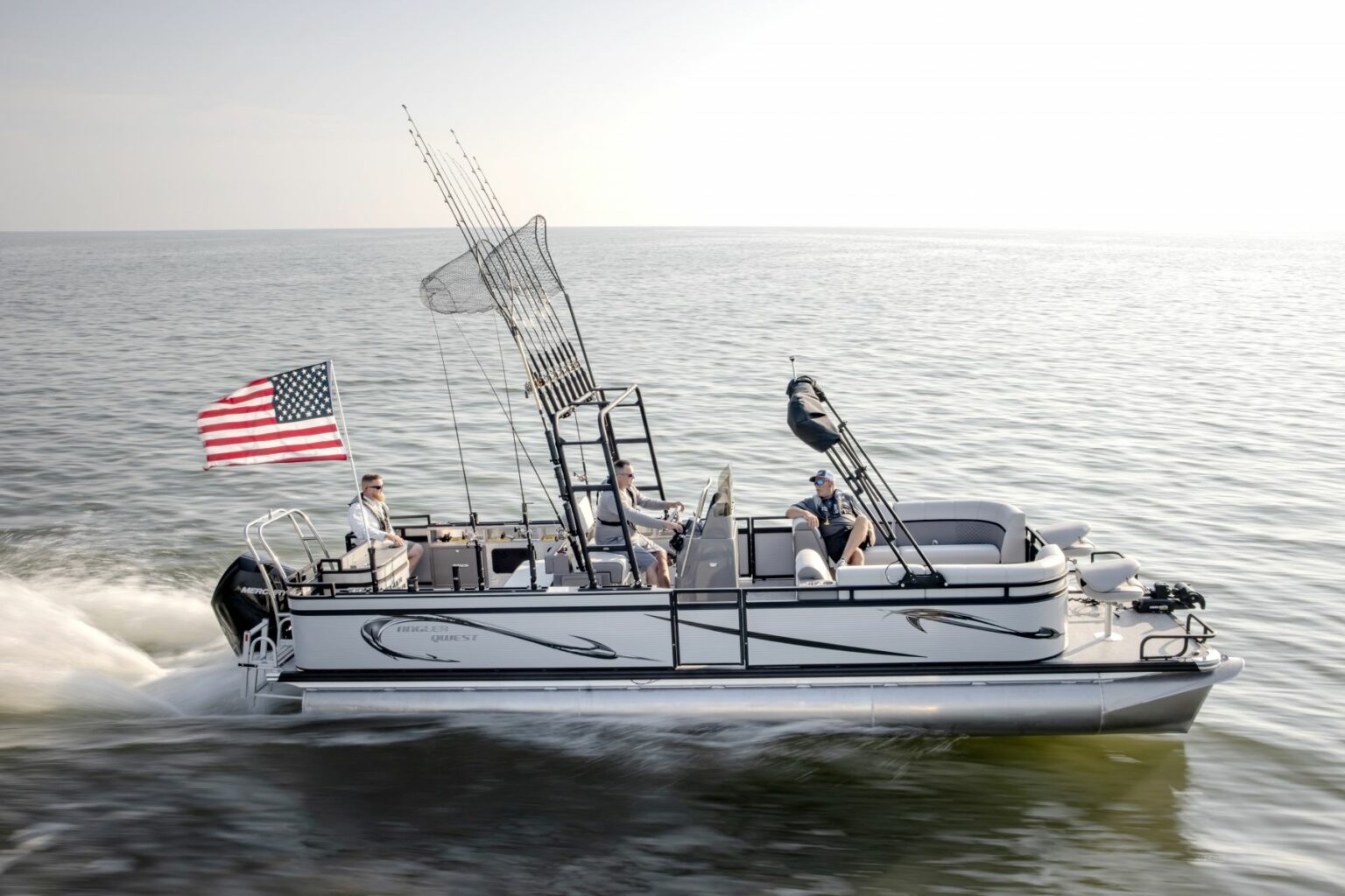 Royalty-free image - A pontoon boat speeds across a calm lake. Three people are on board, with fishing rods and nets visible. An American flag waves at the rear of the boat. The sky above is clear.