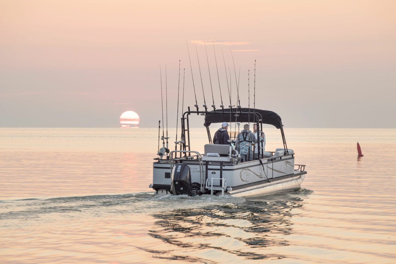 License-free image A small fishing boat with several fishing rods on board moves away from the camera on a calm body of water. The sun is setting on the horizon, casting a warm glow across the sky and water. Two people are visible on the boat, equipped with proper safety equipment, heading towards the sunset.