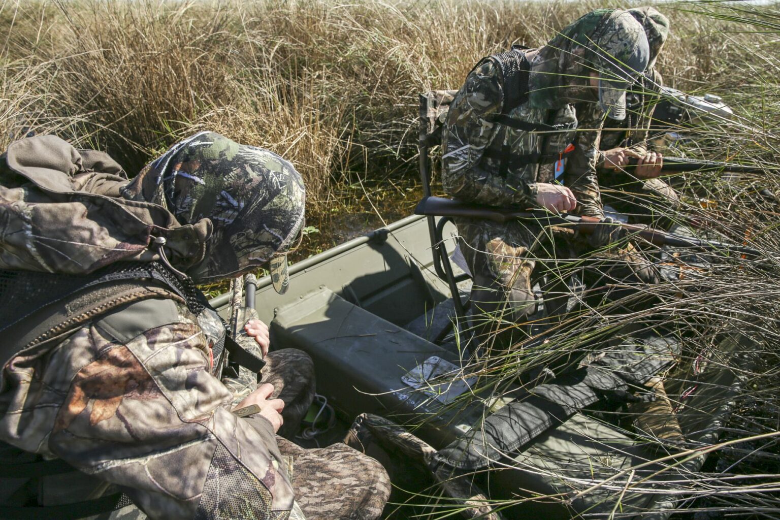 Royalty-free image - Two people in camouflage clothing sit in a small boat, surrounded by tall grass and reeds. They hold shotguns and appear focused on their surroundings, possibly preparing for hunting in a marsh-like environment. Hunting from a jon boat in the marsh along the St. Johns River near Christmas, Florida.