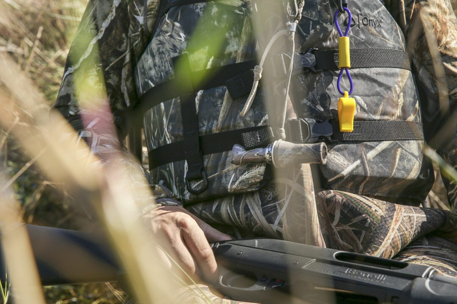 Royalty-free image - A person wearing a camouflage jacket and holding a black shotgun is partially obscured by tall grass. They have whistles attached to their jacket. The setting appears to be an outdoor, natural environment. Hunting from a jon boat in the marsh along the St. Johns River near Christmas, Florida.