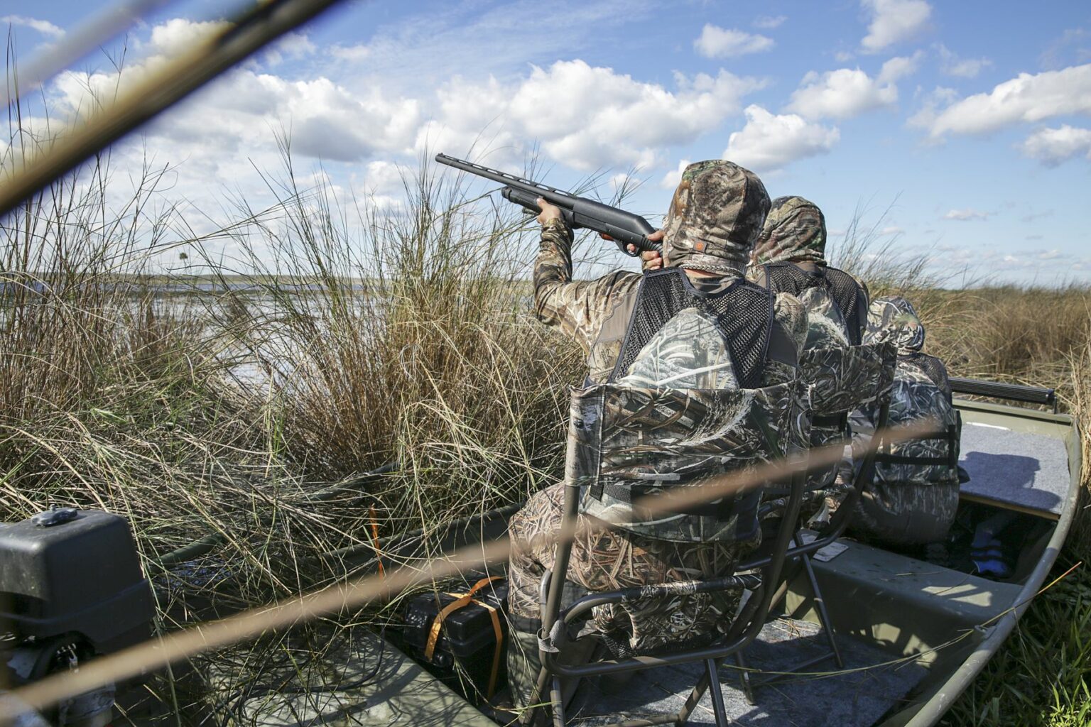 Royalty-free image - Two hunters in camouflage gear sit in a jon boat among tall reeds, aiming shotguns into the sky. The scene unfolds in a marshy area under a partly cloudy blue sky.