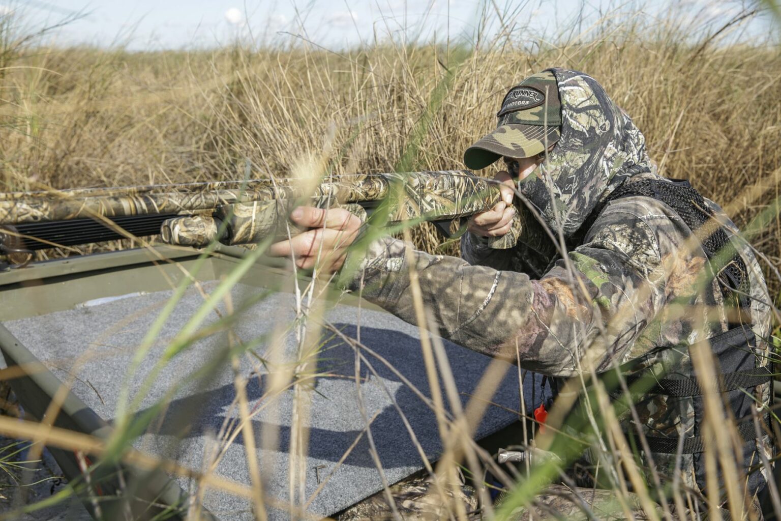 Royalty-free image - A person in camouflage clothing and a cap aims a shotgun while crouching in tall grass near the edge of a wetland. Partially concealed by the surroundings, they look ready to hunt, perhaps after stepping out of their nearby jon boat.