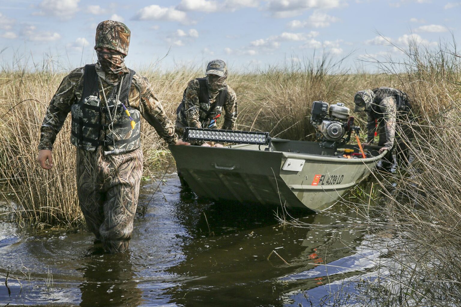 Royalty-free image - Three people in camouflage gear pull a small jon boat through a shallow waterway surrounded by tall grasses. The sky is partly cloudy, enhancing the marshy, wetland atmosphere where the scene unfolds.