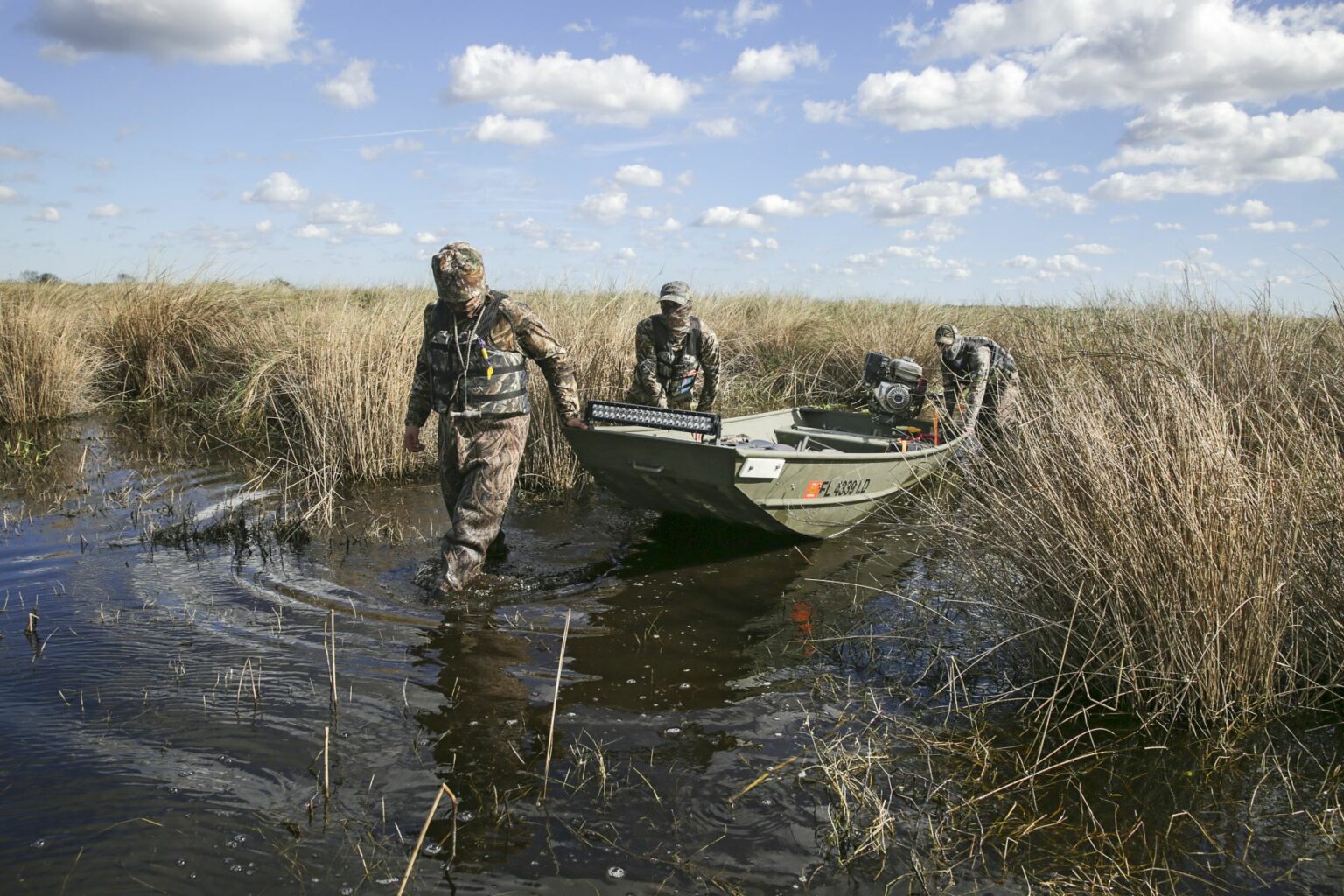 Royalty-free image - Three people in camouflage gear pull a small jon boat through a marshy area. The sky is partly cloudy, and tall grasses surround them as they wade through the shallow water.