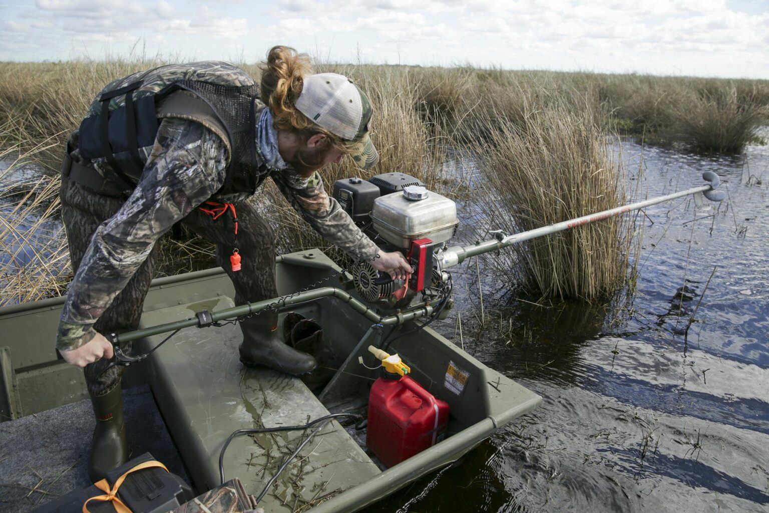 Royalty-free image - A person in camouflage clothing and a cap is standing on a small boat in a marshland, operating a long-handled pole tool near the water. Tall grasses surround the area under a partly cloudy sky. Hunting from a jon boat in the marsh along the St. Johns River near Christmas, Florida.