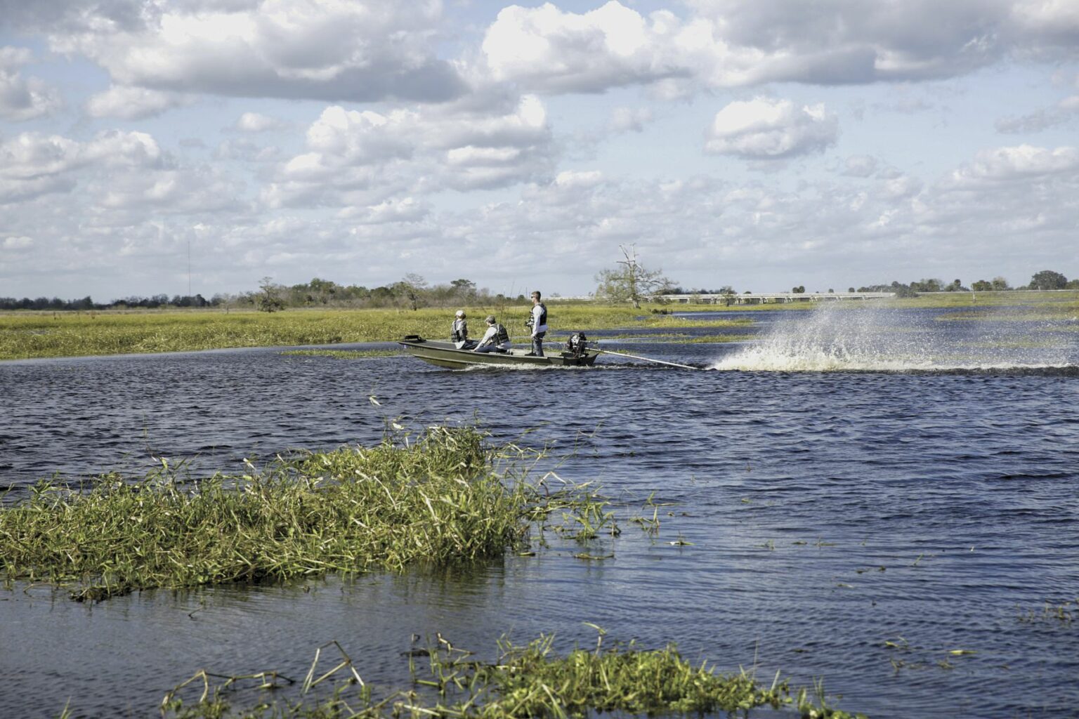 Royalty-free image - A small jon boat speeds across a lake, leaving a spray of water behind. Three people are on board, surrounded by vast stretches of water and patches of grass under a cloudy sky.