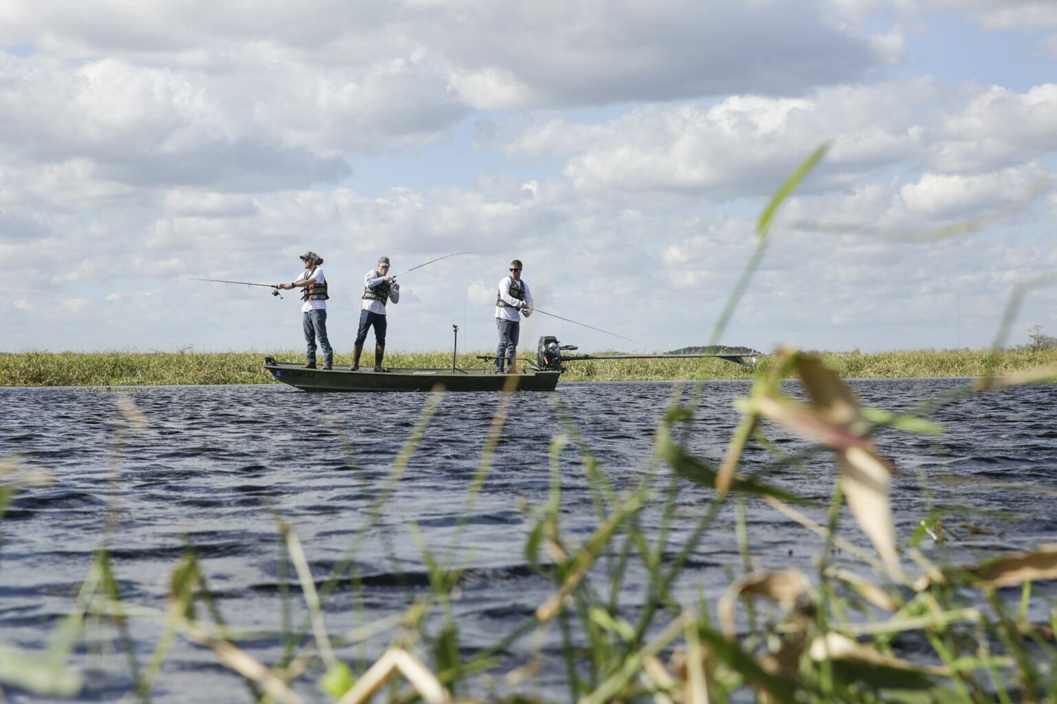 Royalty-free image - Three people are fishing on a small jon boat in a wide, calm lake. The sky is partly cloudy, and tall grass is visible in the foreground, framing the serene scene.