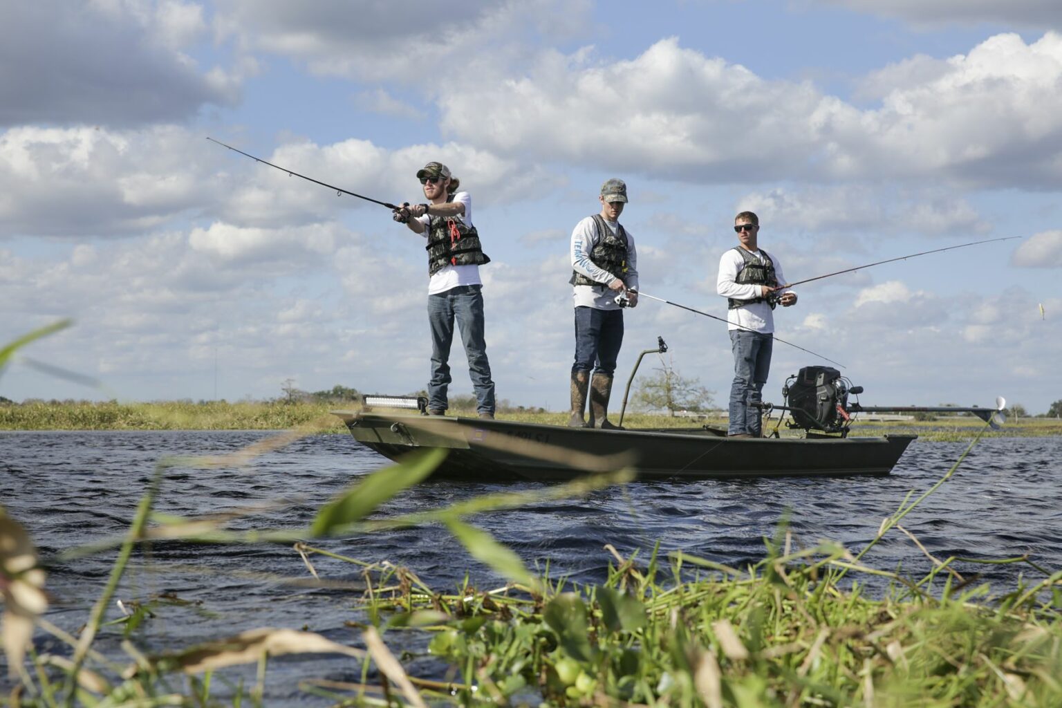 Royalty-free image - Three people are fishing from a small jon boat in a grassy lake under a partly cloudy sky. They are wearing casual clothing and hats, each holding fishing rods, with the background showing open water and distant trees.