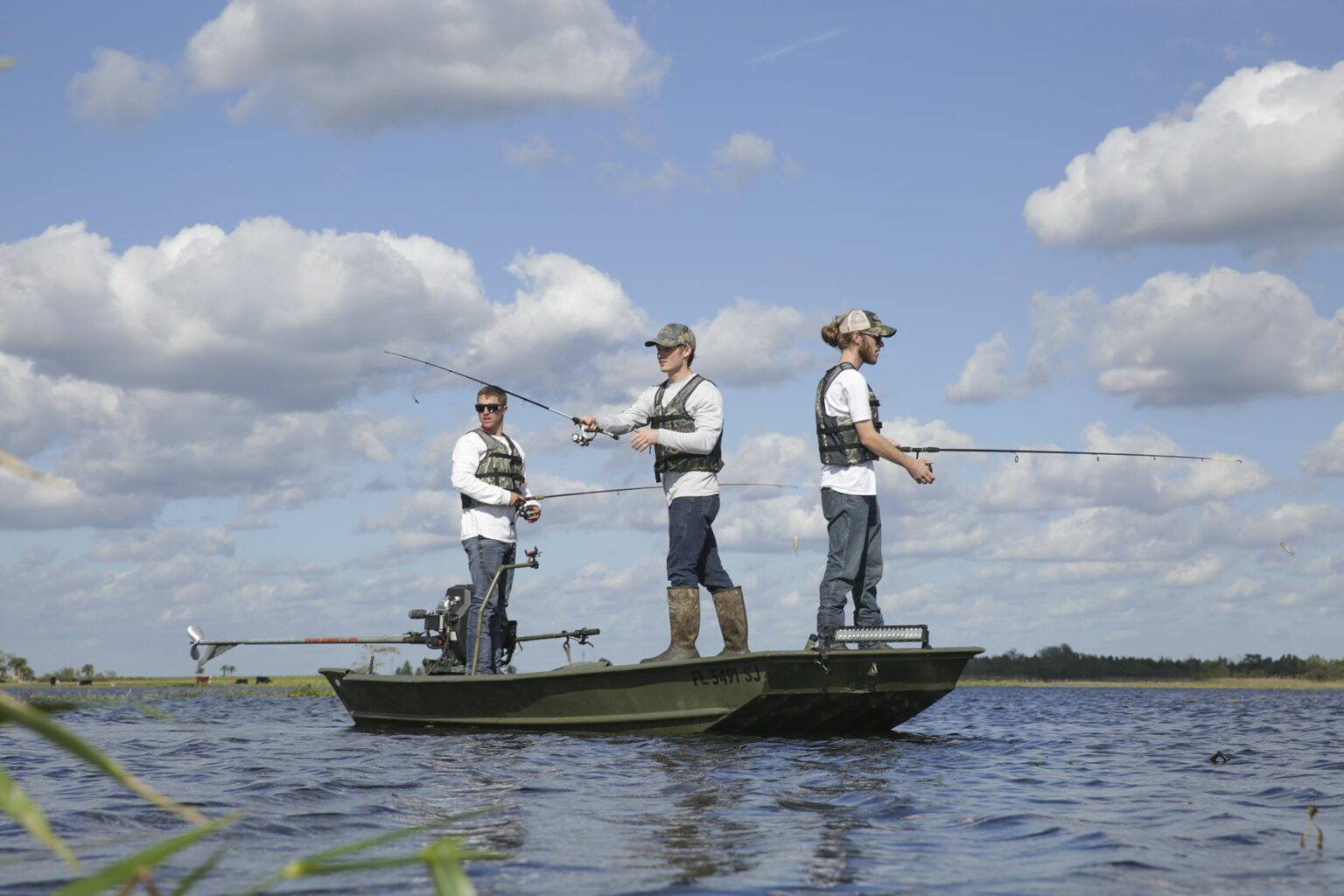 Royalty-free image - Three people in casual attire are fishing on a jon boat in a large body of water under a partly cloudy sky. They each hold fishing rods, casting their lines into the calm water. The scene is tranquil with distant land visible.