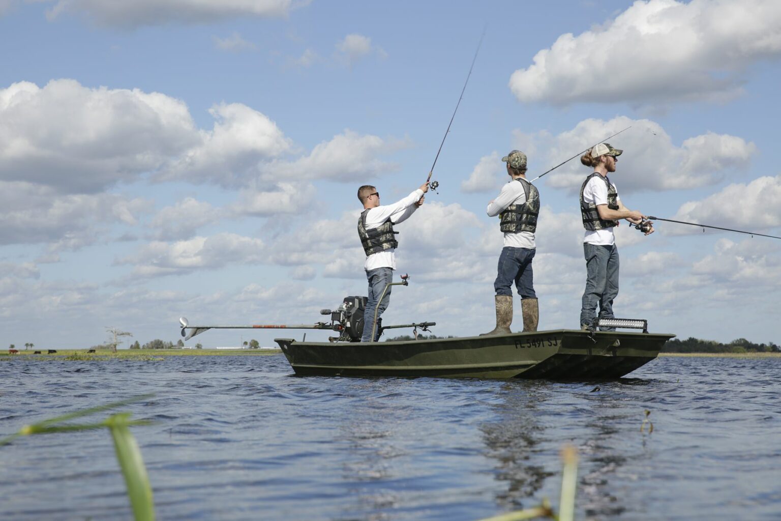 Royalty-free image - Three people in life vests fish from a small jon boat on a calm lake. Under a partly cloudy sky, they&#039;re surrounded by open water with distant shorelines visible. One person casts a line while the others hold fishing rods, enjoying the serene day.