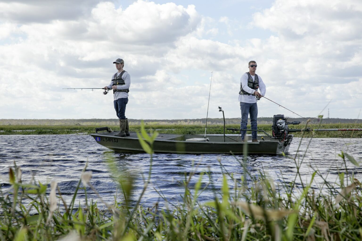 Royalty-free image - Two people fishing on a jon boat in a lake surrounded by grassy banks. They are wearing life jackets, and it is a partly cloudy day. The water is calm, with an open sky and distant shoreline providing the perfect backdrop for their peaceful outing.