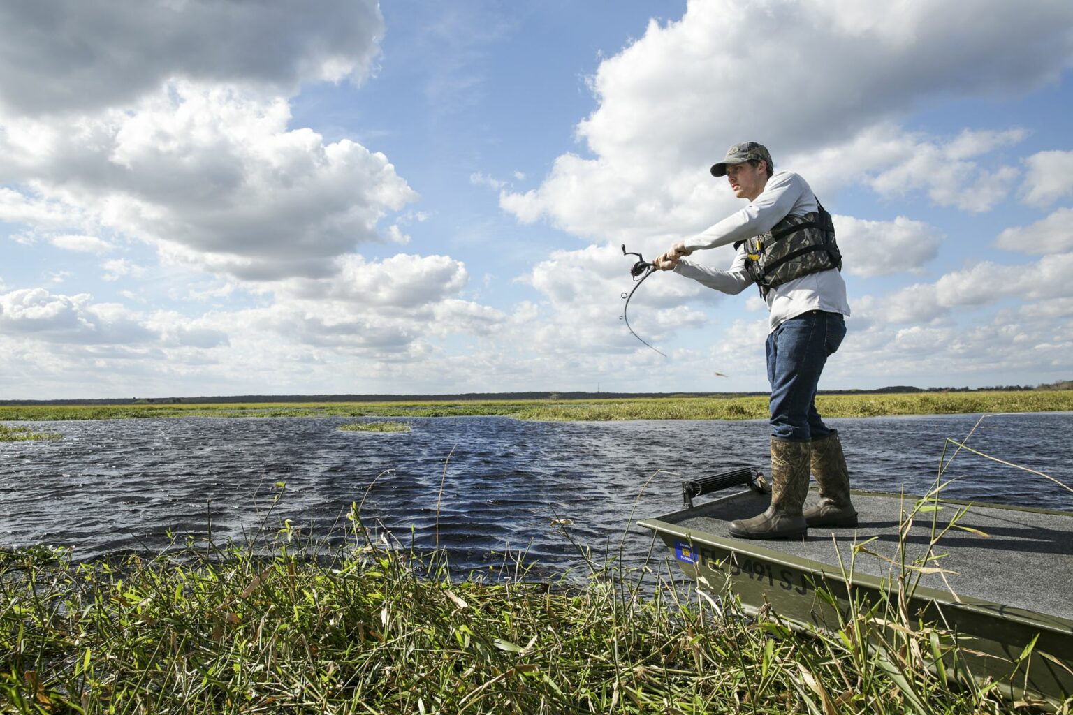 Royalty-free image - A person wearing a cap and life vest casts a fishing line from their jon boat into the water under a partly cloudy sky. They stand amidst grass and reeds, looking focused, with a wide expanse of water and marsh stretching out in the background.