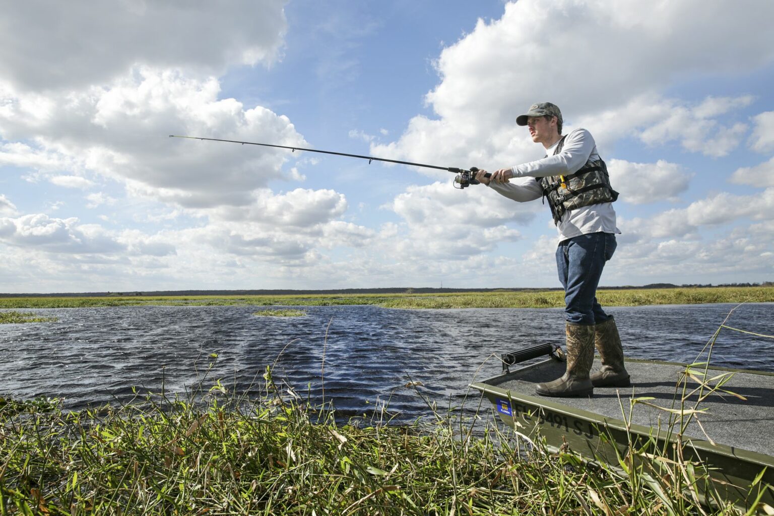 Royalty-free image - A person in casual outdoor attire is standing on a jon boat, casting a fishing line into a lake surrounded by grass and reeds. The sky is partly cloudy, enhancing the peacefulness of this serene outdoor fishing experience.