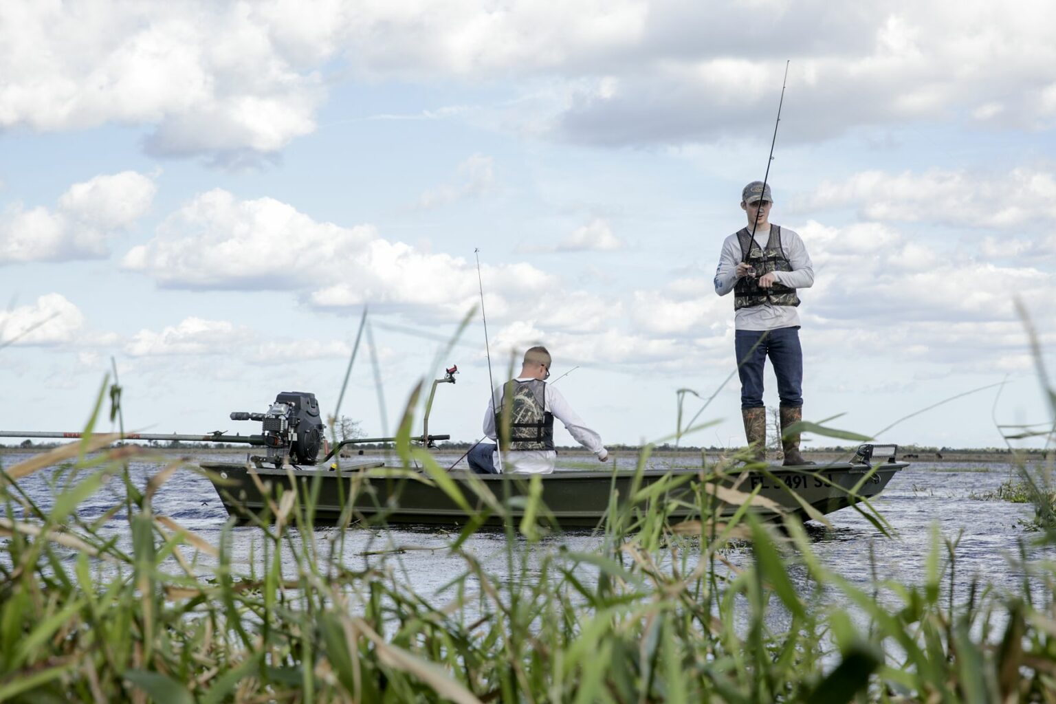 Royalty-free image - Two people fishing from a small jon boat on a river or lake. One is standing with a fishing rod, while the other is seated. The scene is set against a cloudy sky with tall grasses in the foreground.