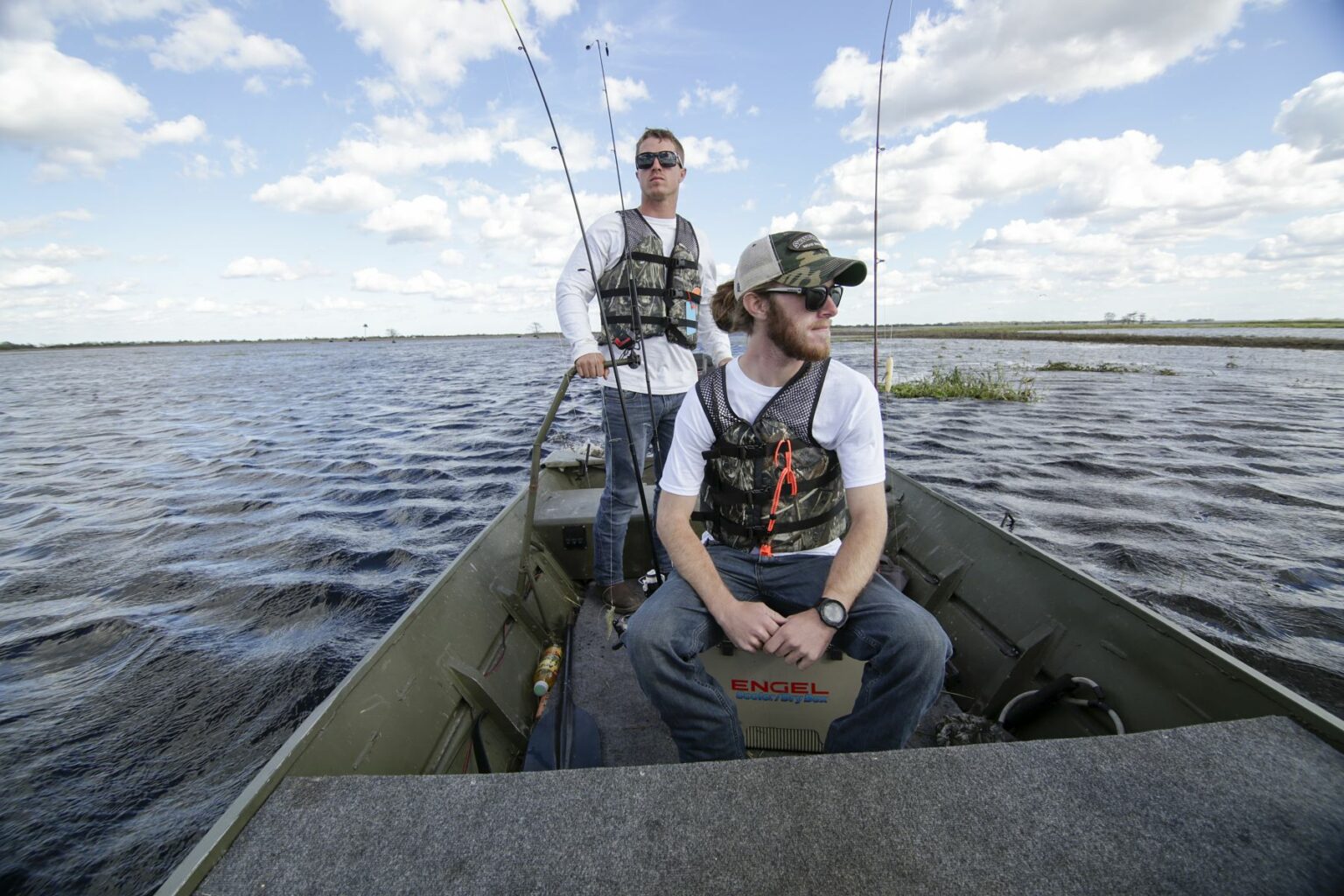 Royalty-free image - Two men in casual attire and life vests are on a jon boat in a large body of water, fishing rods nearby. One sits on a cooler, the other stands. The sky is filled with scattered clouds, suggesting a pleasant day for fishing.