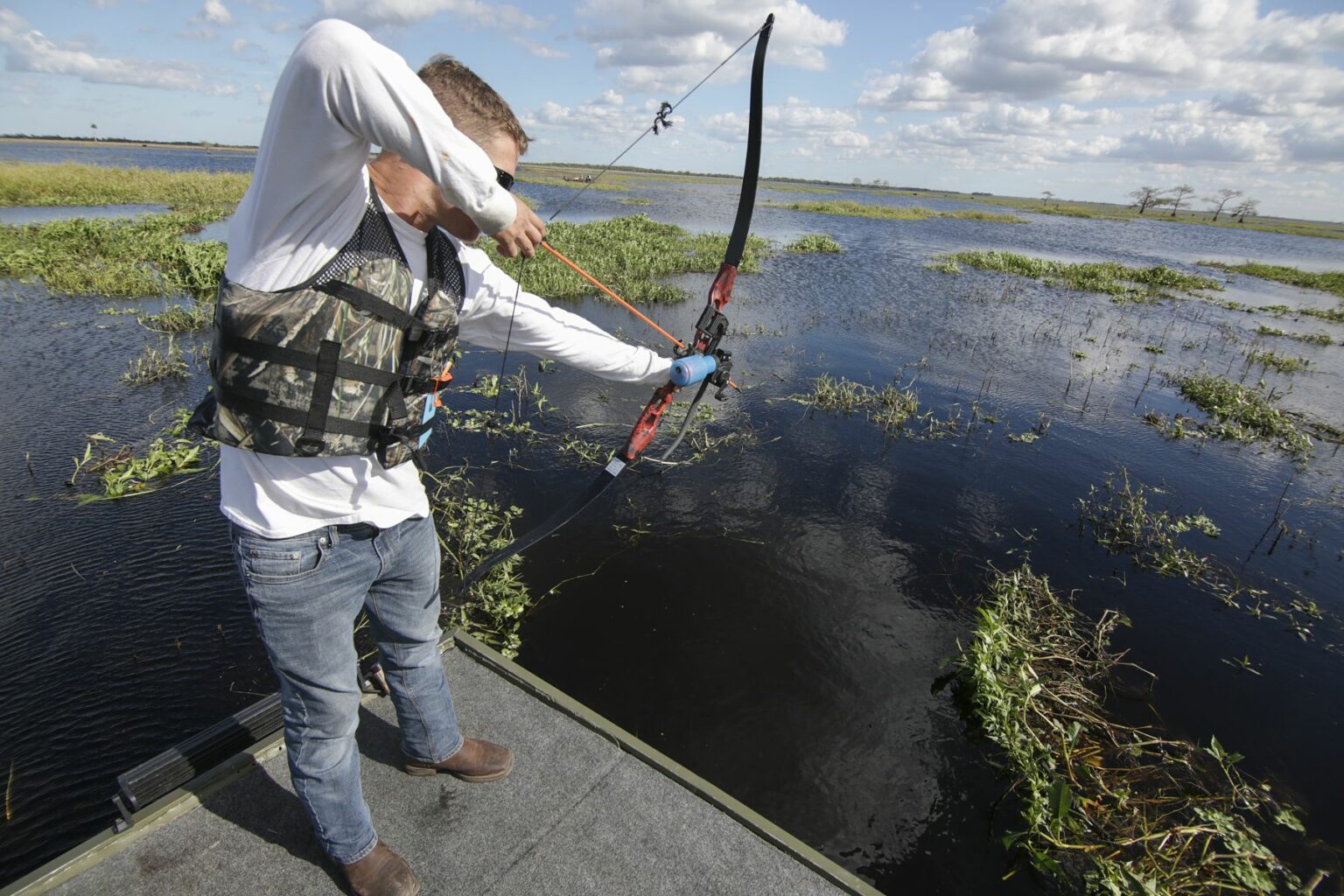 Royalty-free image - A person in a camouflage vest and jeans uses a bow to fish from a jon boat on a body of water under a partly cloudy sky. The water is dotted with small green plants.