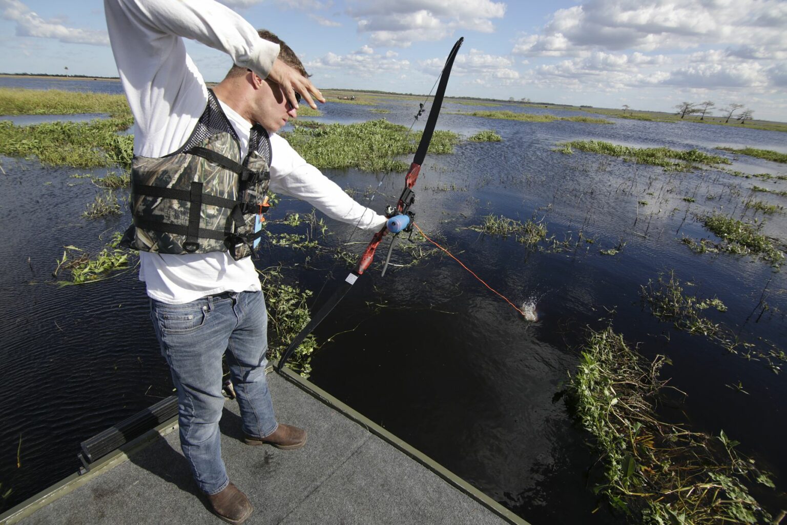 Royalty-free image - A person on a jon boat, wearing a camouflage life vest, skillfully uses a bow to fish in the vast, watery landscape with scattered vegetation under a partly cloudy sky.